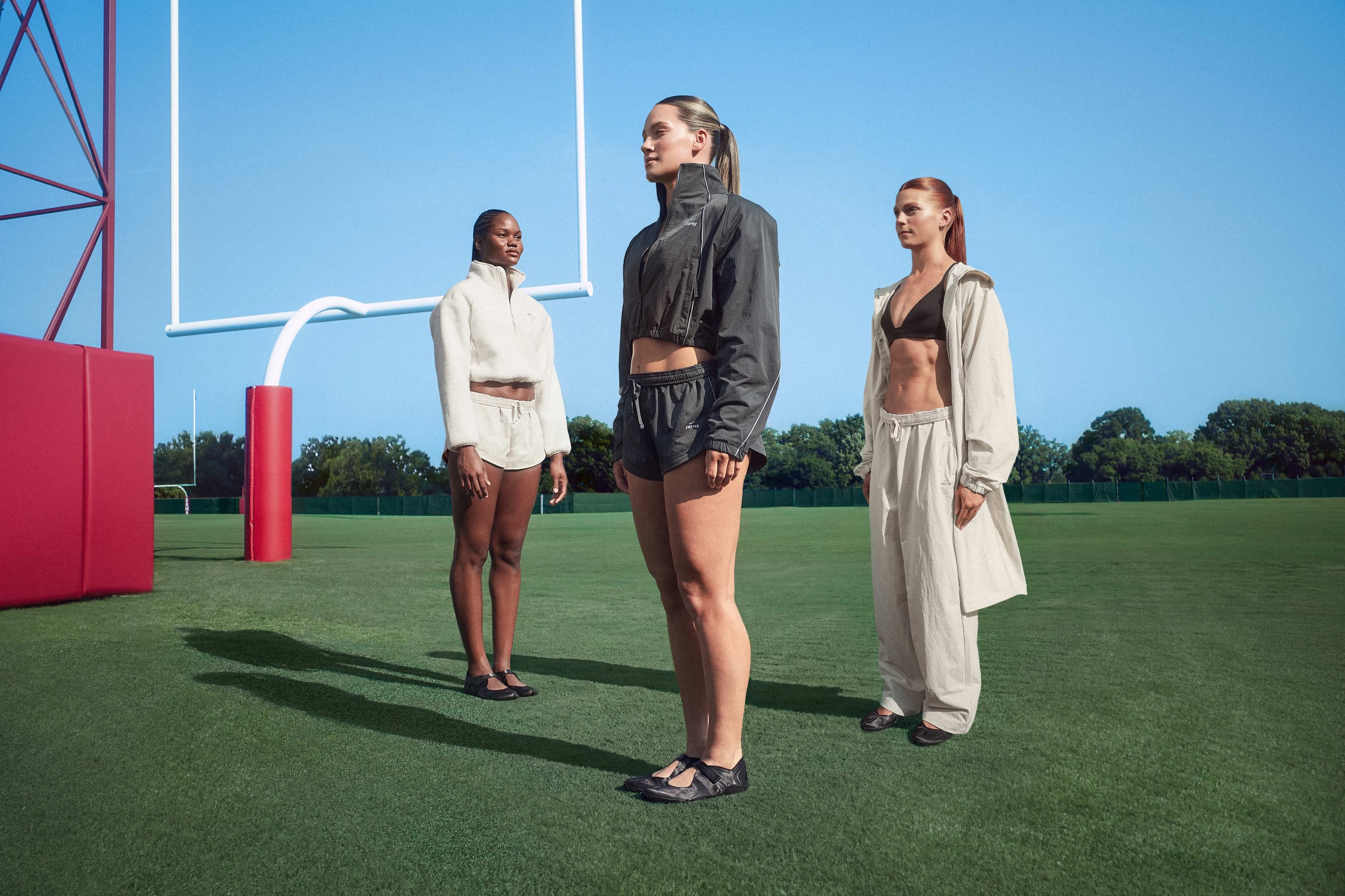 Three women stand on a football field, dressed in athletic wear. A goalpost is visible in the background under a clear blue sky.