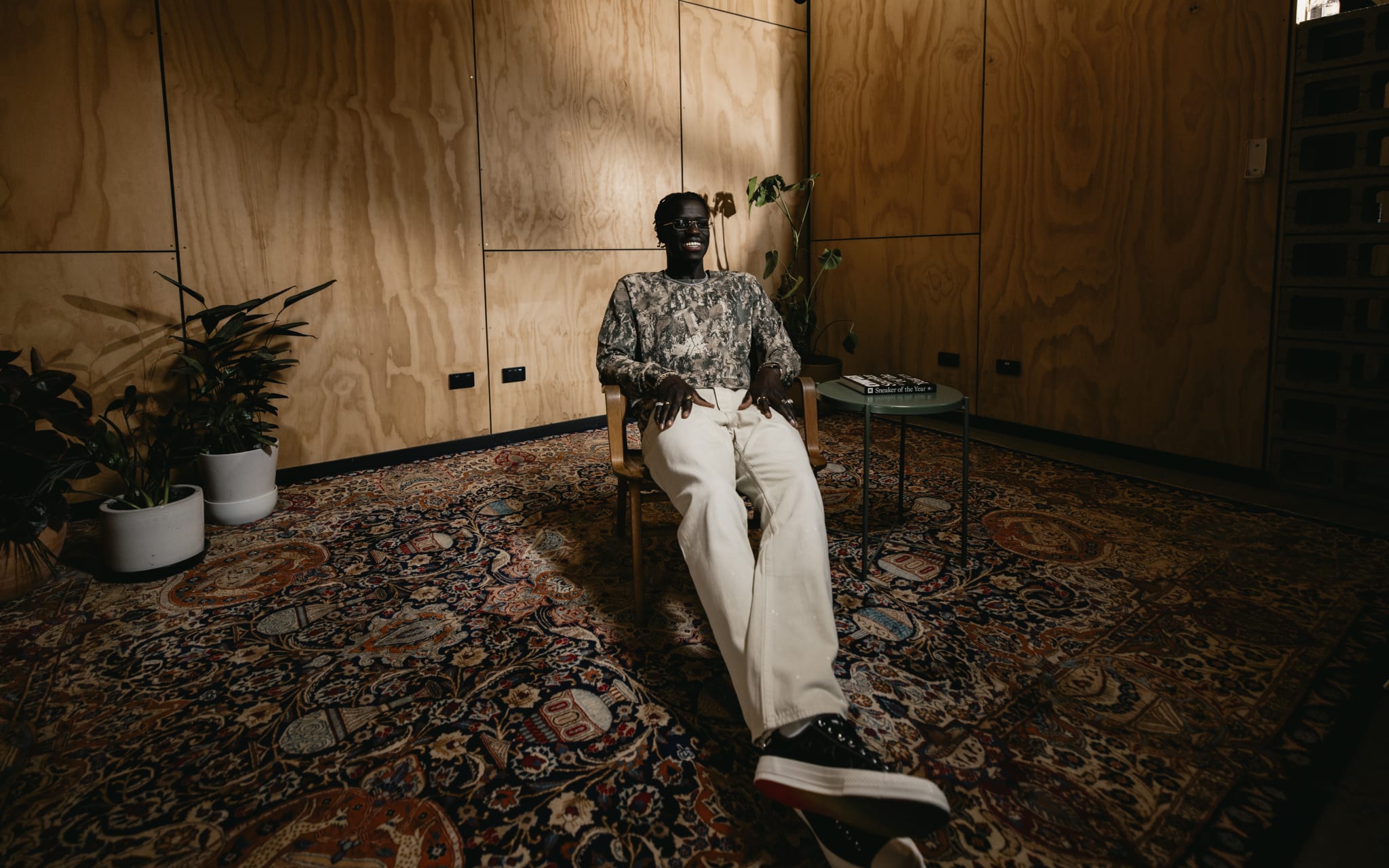 A person sits in a wooden room with a patterned rug, surrounded by plants, under a microphone, creating a cozy atmosphere.