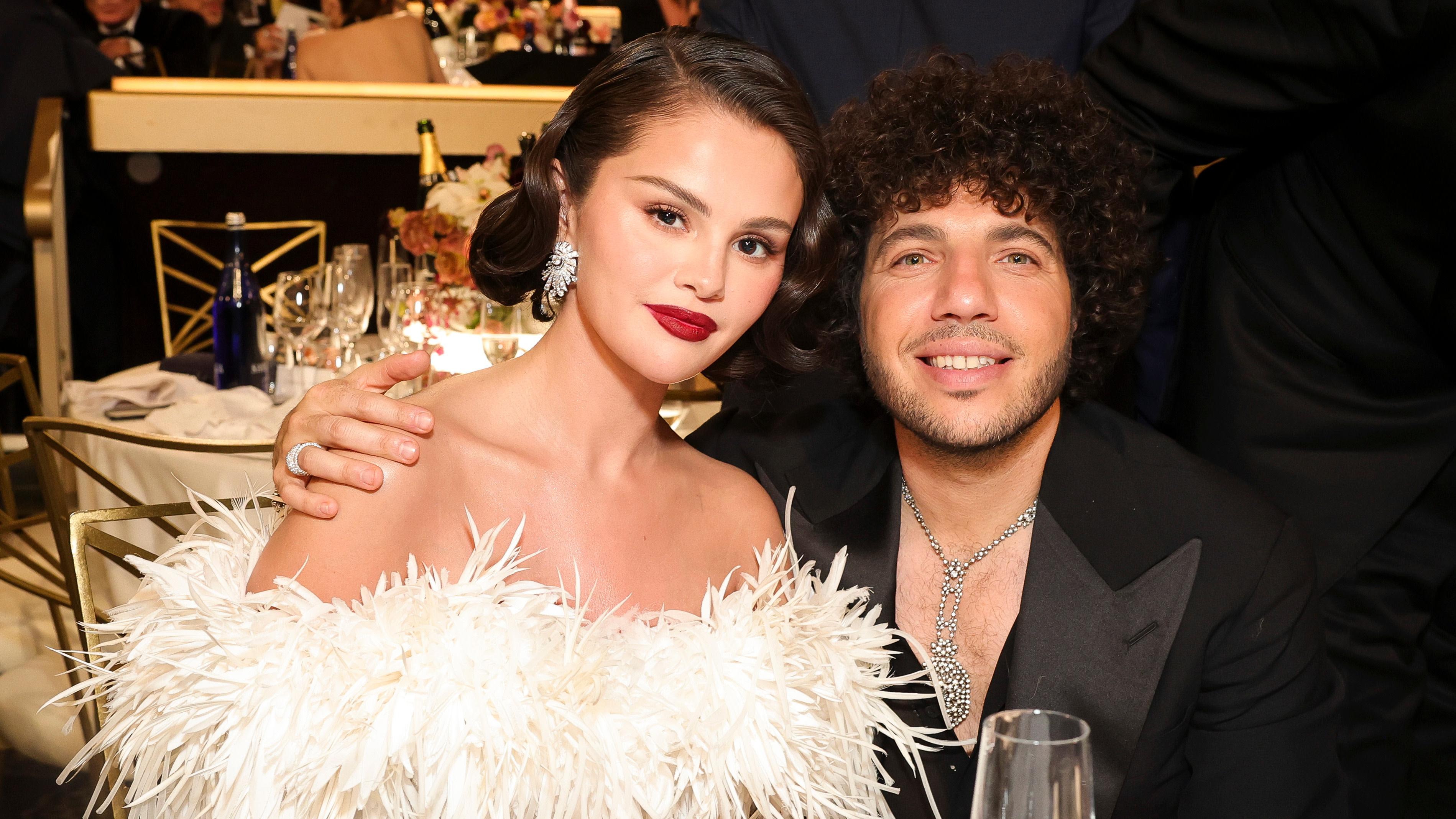 Selena Gomez in a white feathered dress sits beside Benny Blanco in a black suit at a formal event.