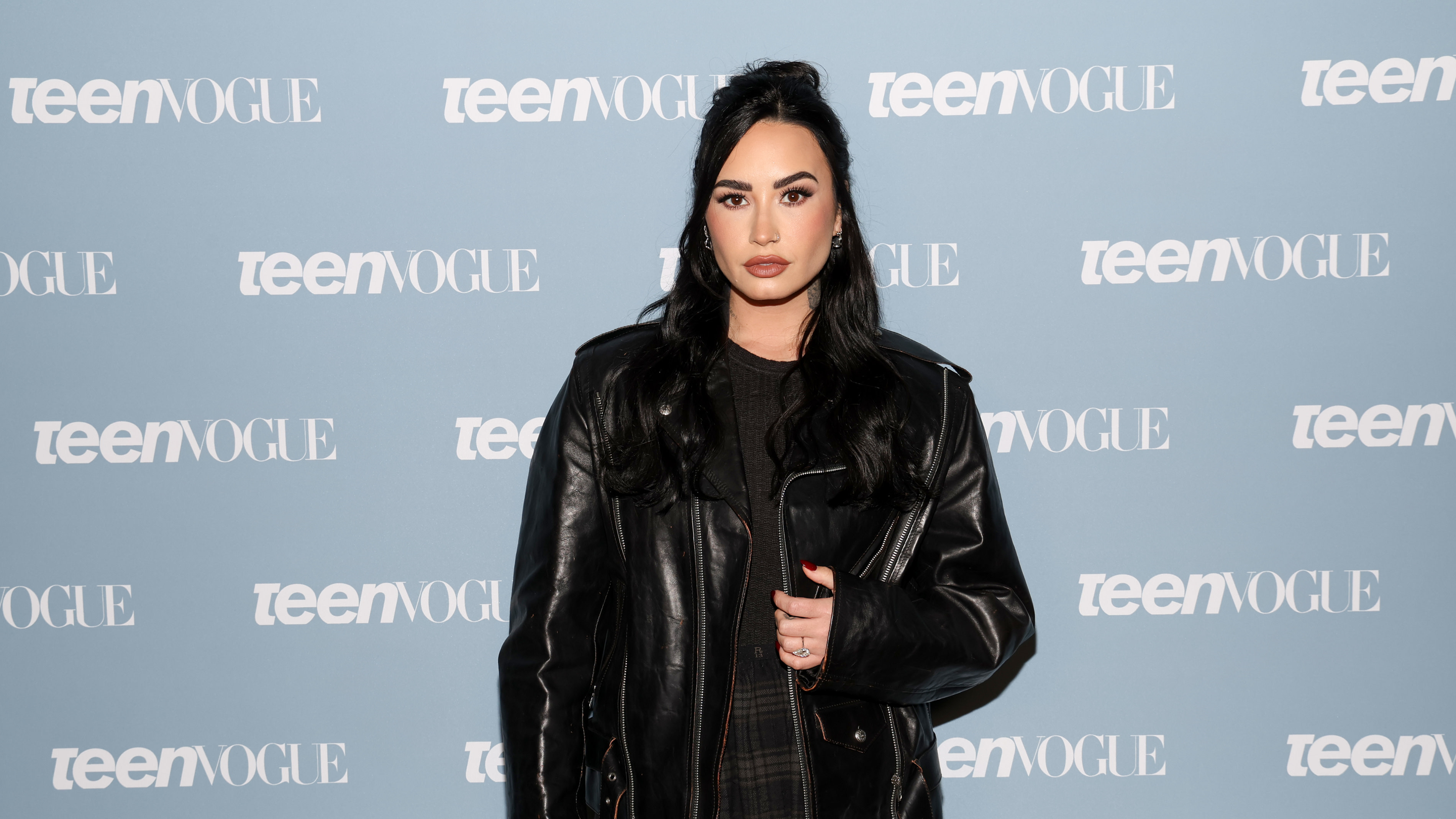 Demi Lovato at a Teen Vogue event, wearing a black leather jacket, with long dark hair and a neutral expression.