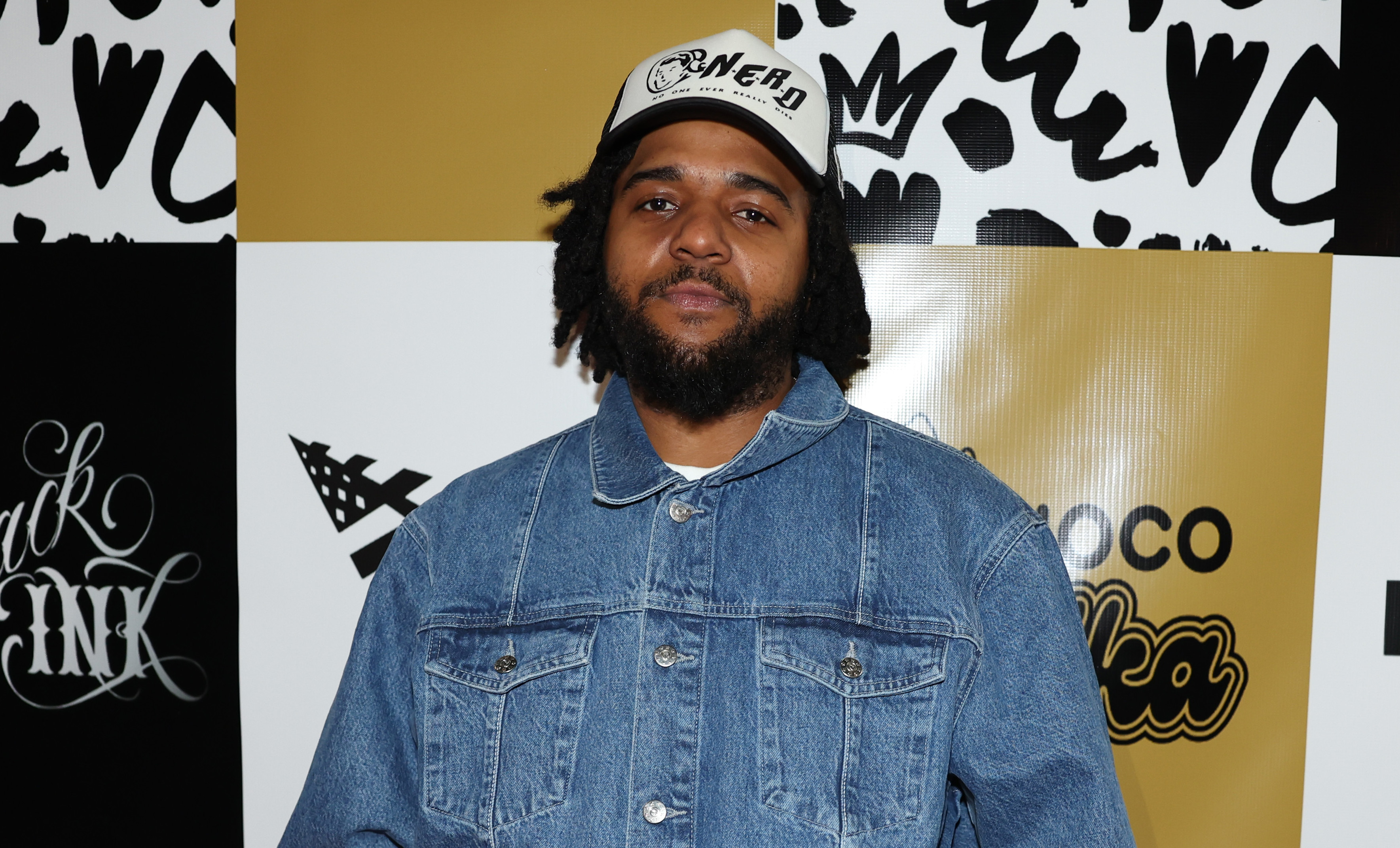 CJ Wallace in a denim outfit and cap stands in front of a backdrop with logos including "The Biggie Experience" and "Black Ink."