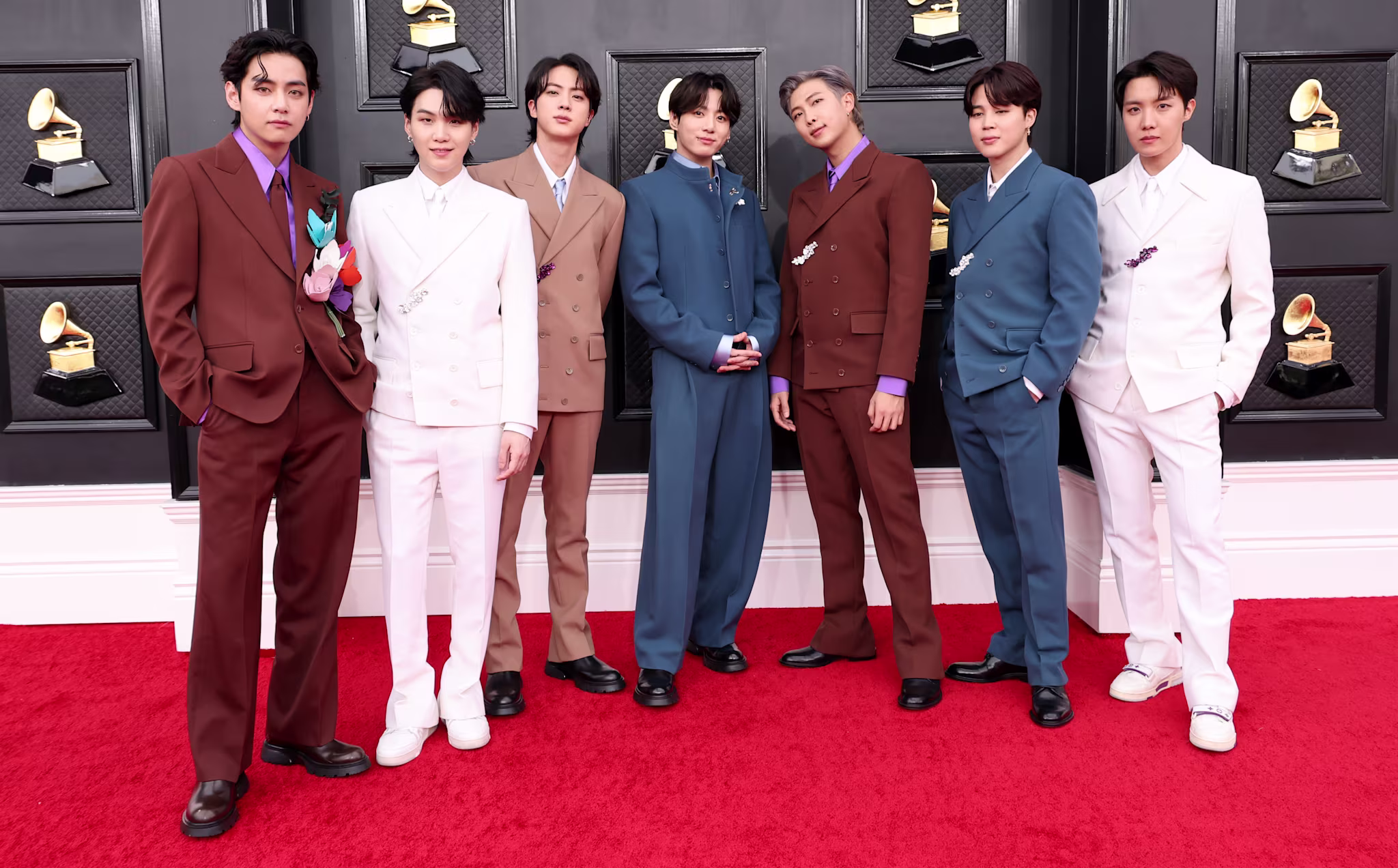 BTS members on the red carpet at the Grammy Awards, wearing stylish suits in various colors, standing in front of a backdrop with Grammy logos.