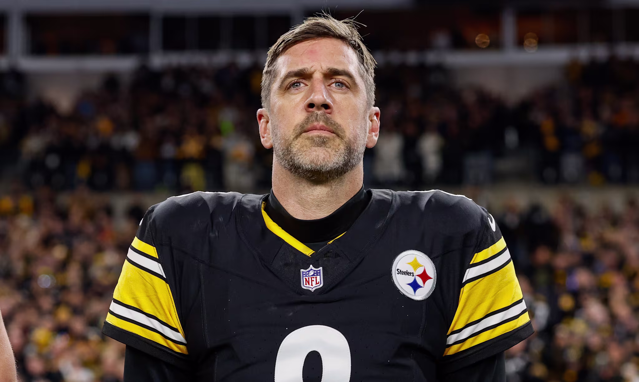 Aaron Rodgers wearing a Pittsburgh Steelers jersey with number 8 stands on the field, looking focused.