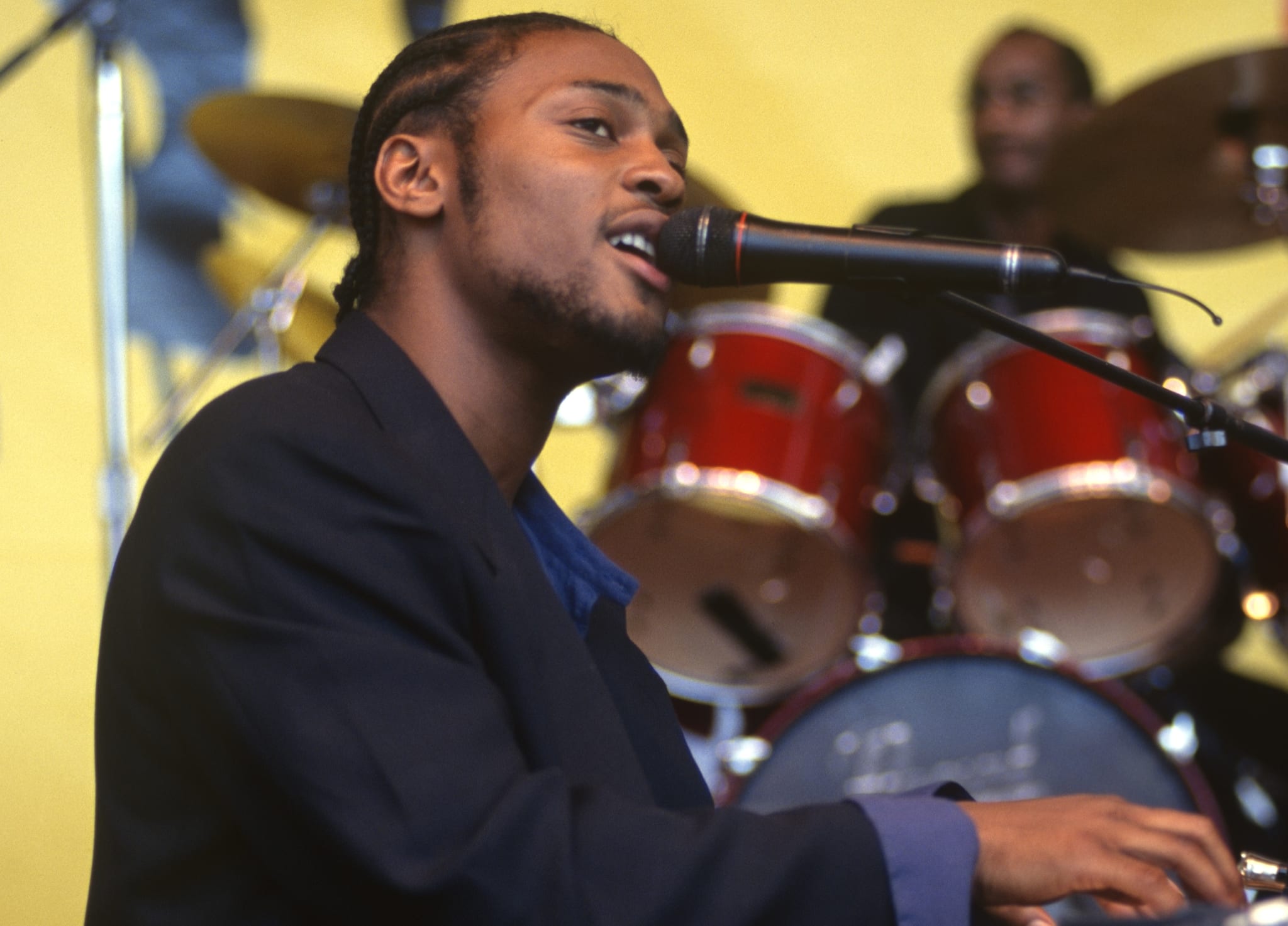 A musician with braided hair sings into a microphone while playing the keyboard, with a drummer in the background.