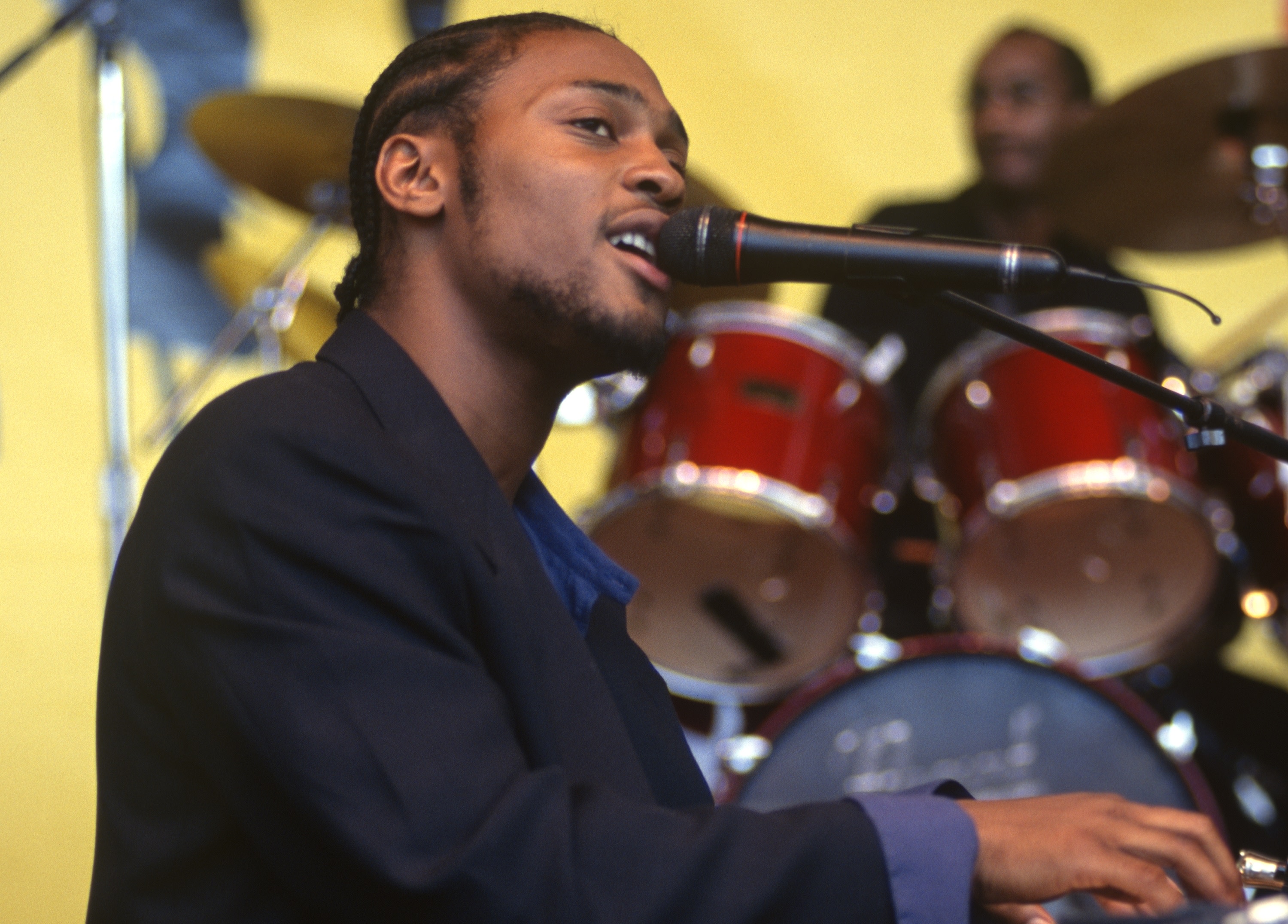 A musician with braided hair sings into a microphone while playing the keyboard, with a drummer in the background.