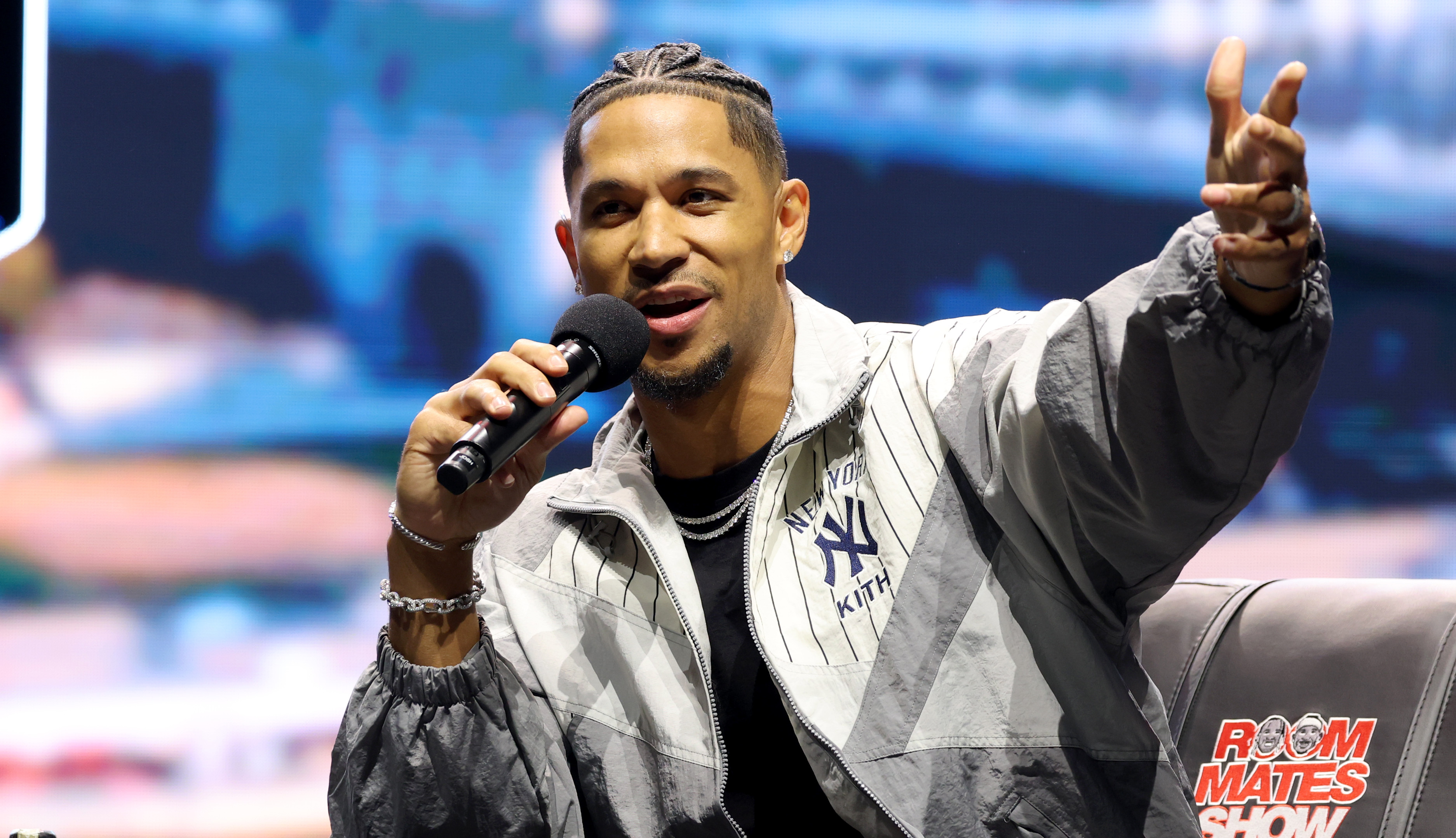 Josh Hart, with braided hair, speaks into a microphone on stage, sitting on a black chair with "Roommates Show" written on it. A bottle is on the table.