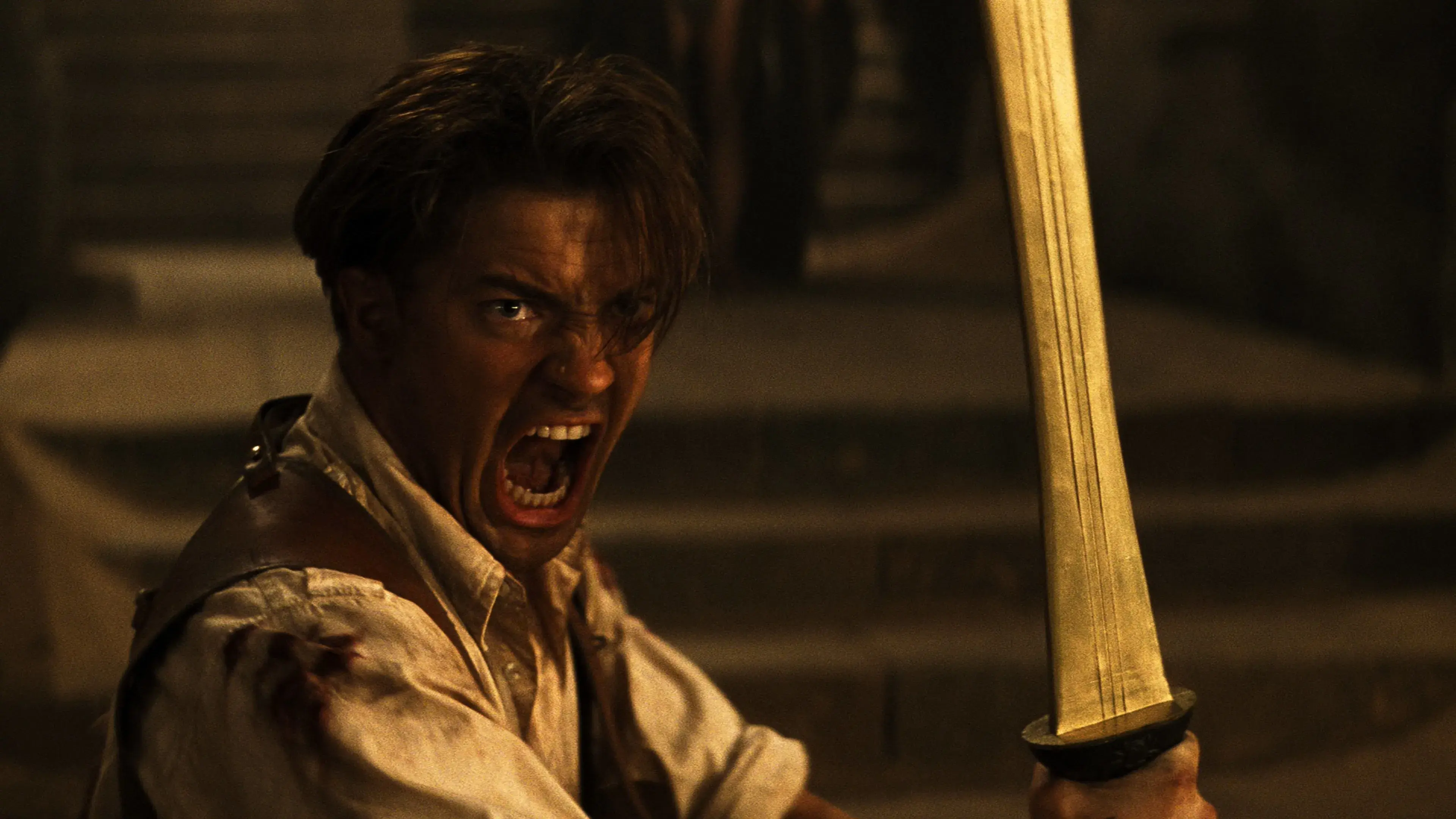 Brendan Fraser in The Mummy in a distressed state, holding a sword, wearing a white shirt with a leather strap, in a dimly lit setting.