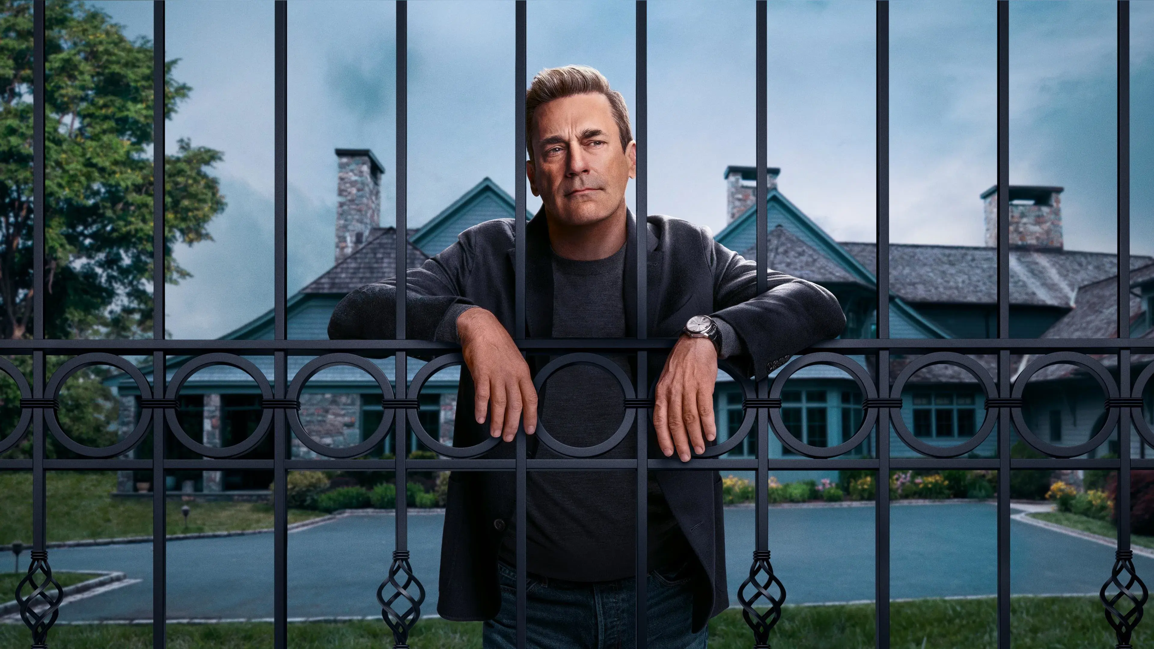 A man stands behind an ornate gate, with a large house and driveway in the background.