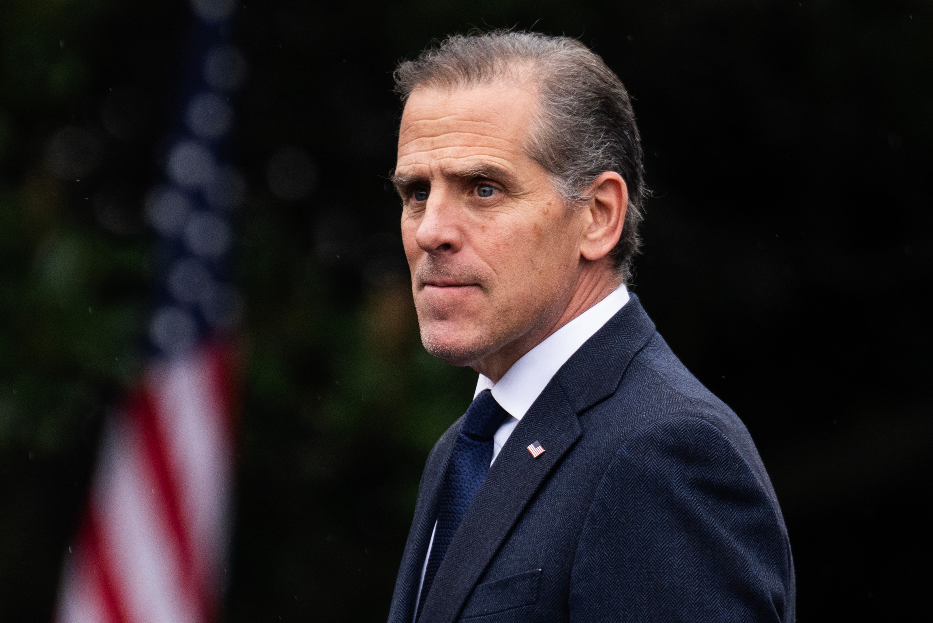 Hunter Biden in a suit stands outdoors with an American flag in the background.