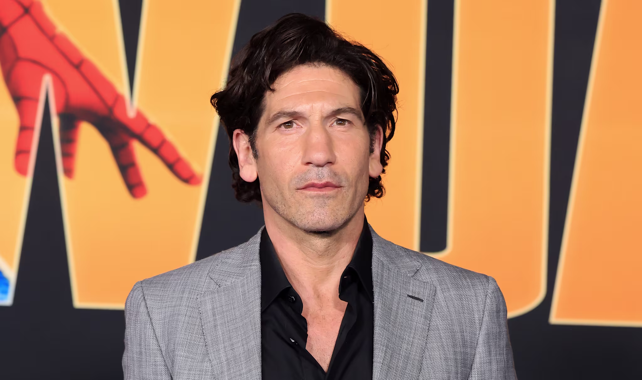 Jon Bernthal in a gray suit and black shirt poses against a backdrop with bold yellow text and a Spider-Man graphic.