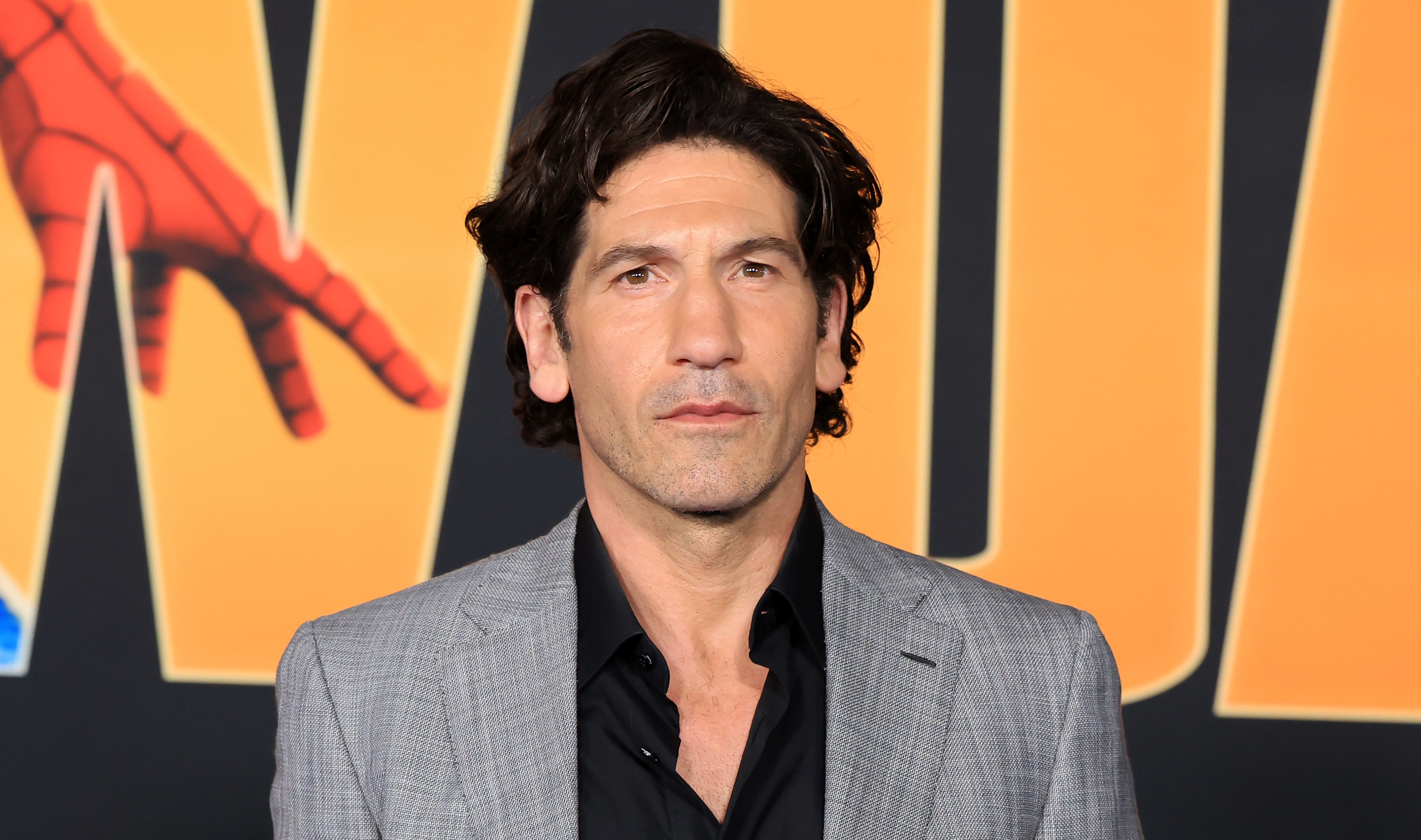 Jon Bernthal in a gray suit and black shirt poses against a backdrop with bold yellow text and a Spider-Man graphic.