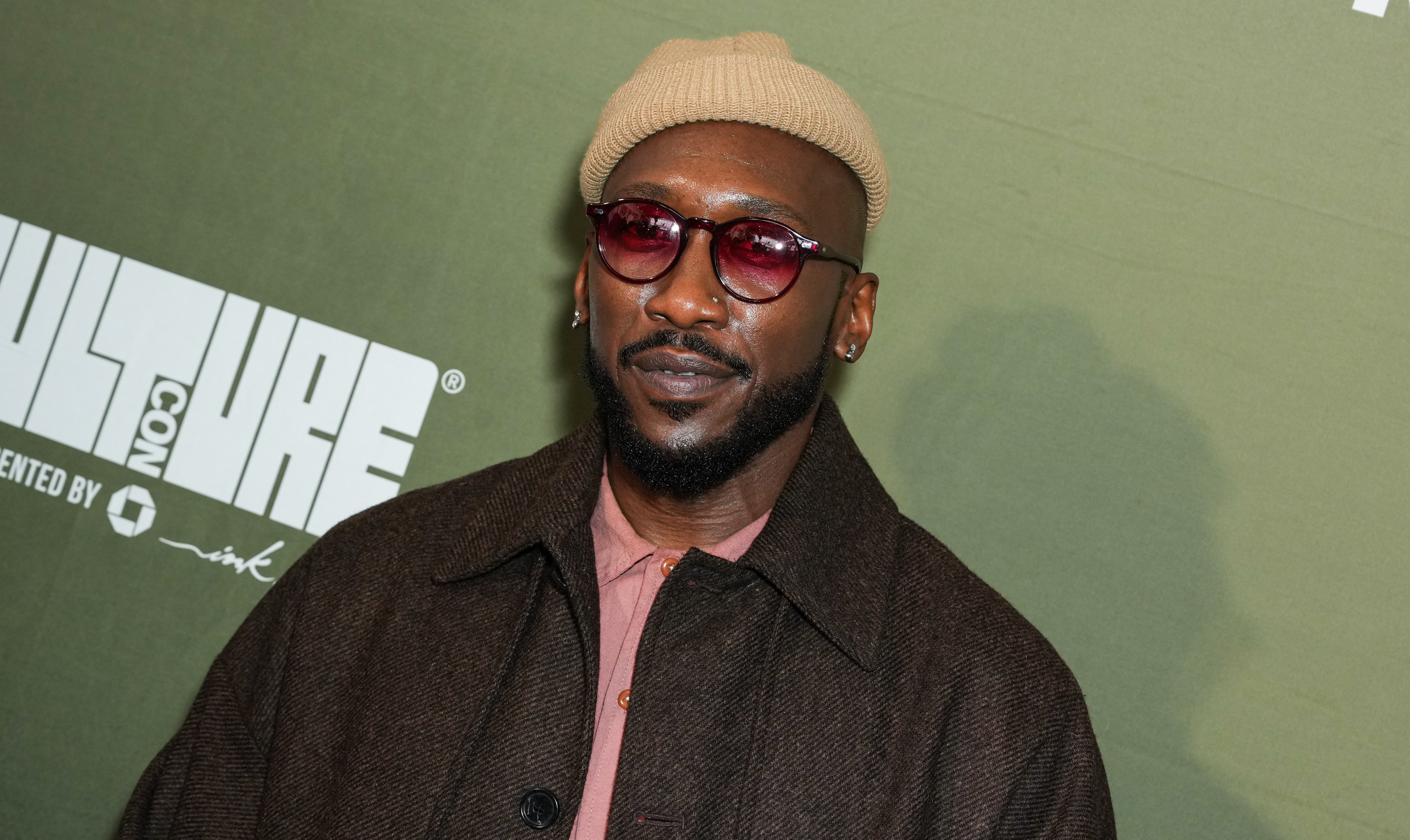 Mahershala Ali wearing a beanie and sunglasses, standing in front of a green backdrop with "Culture Con" text.