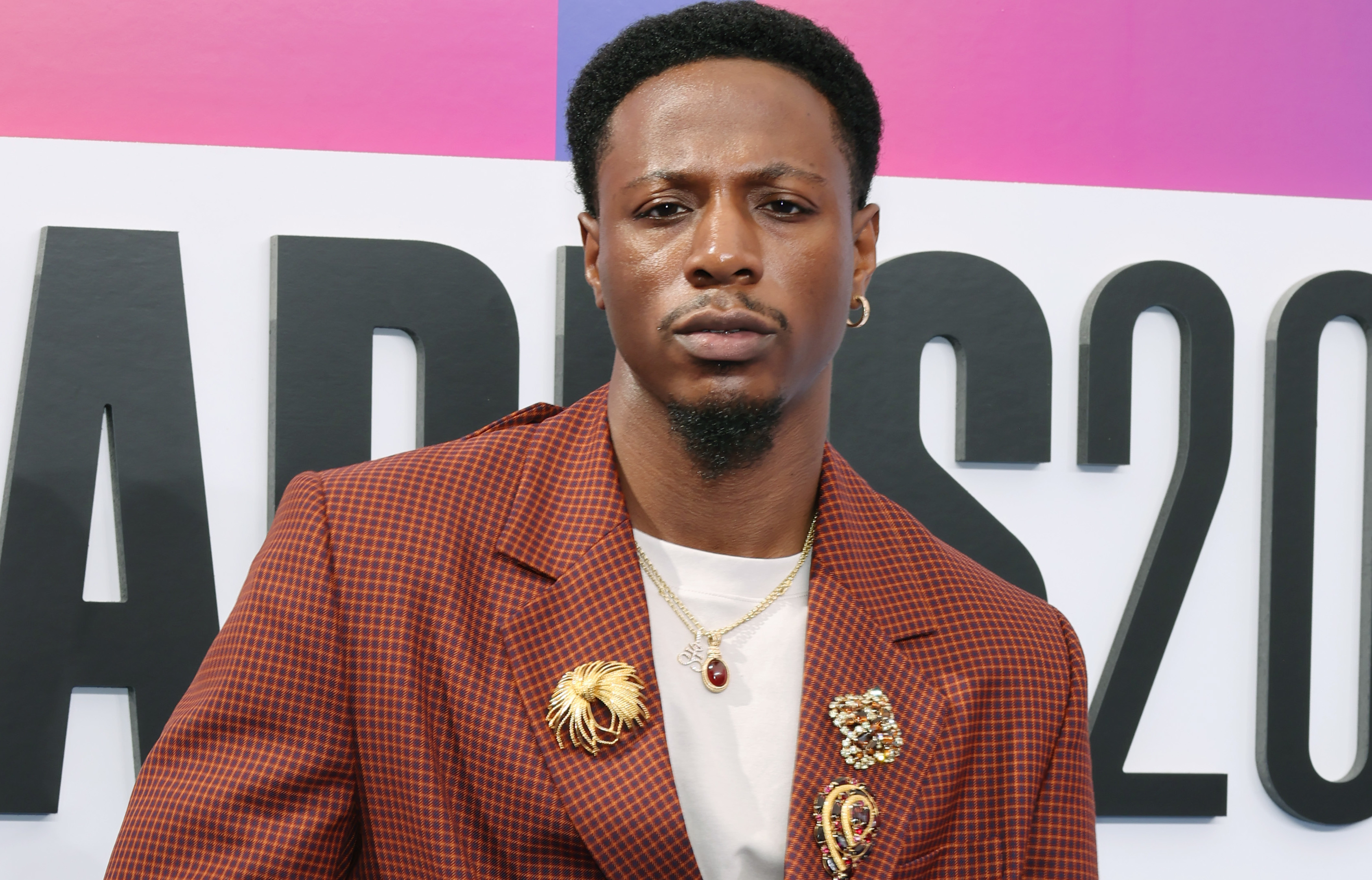 Joey Badass in a red checkered suit with gold accessories, standing in front of an awards backdrop.