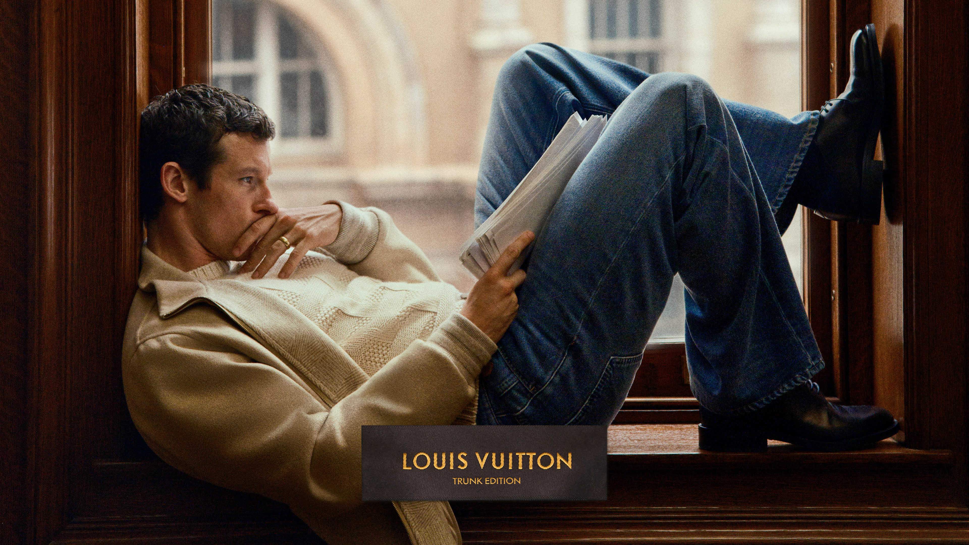 Callum Turner in a cream sweater and jeans sits in a window reading. "LOUIS VUITTON TRUNK EDITION" text is at the bottom.