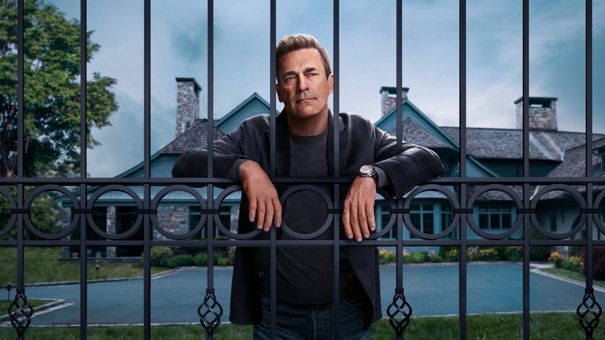 A man stands behind a decorative iron fence in front of a large house with a pool. The sky is overcast, and there are trees in the background.
