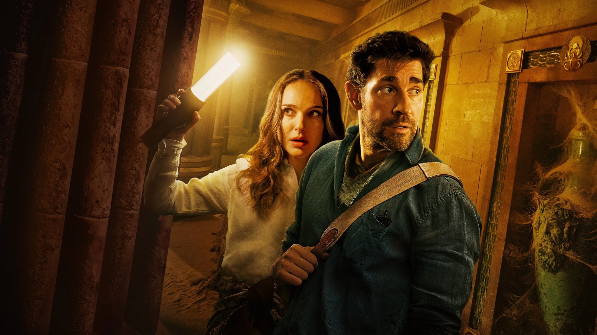 A man and woman in an ancient, dimly lit setting. The woman holds a glowing object, and both appear cautious, looking in opposite directions.