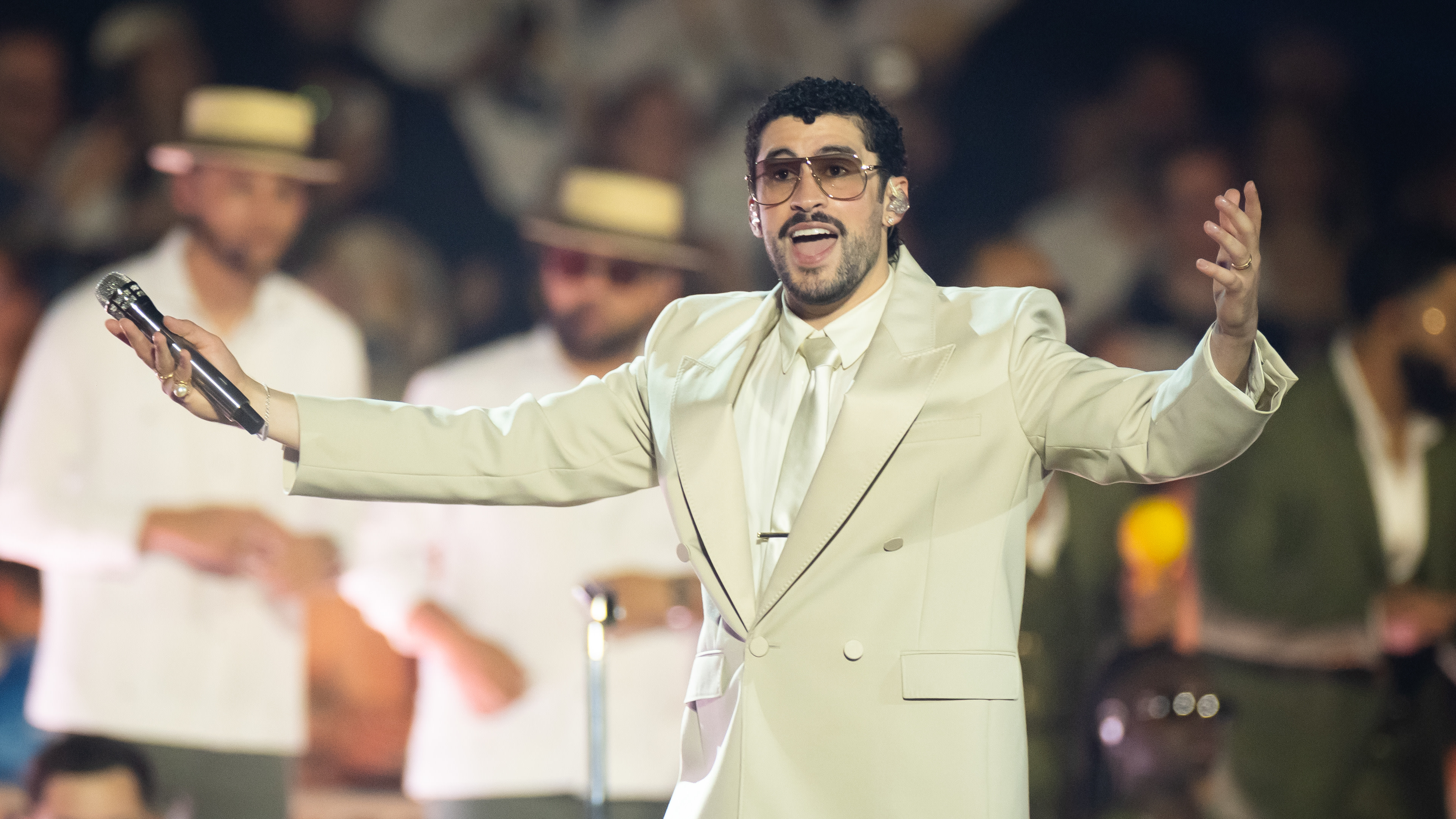 Bad Bunny performing on stage, wearing a beige suit and sunglasses, holding a microphone with arms outstretched.