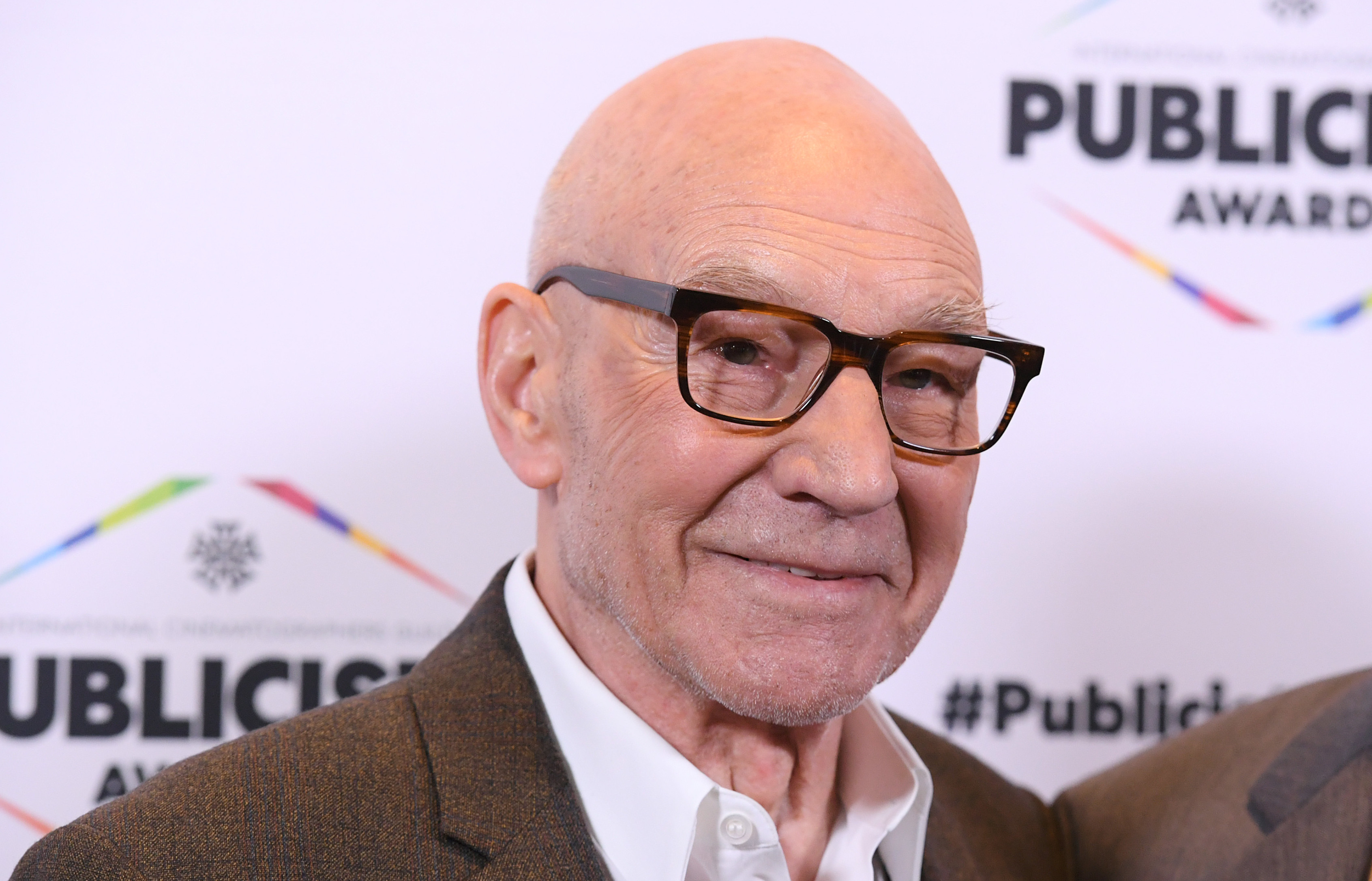 Patrick Stewart at the Publicists Awards, wearing glasses and a suit, smiling at the camera.