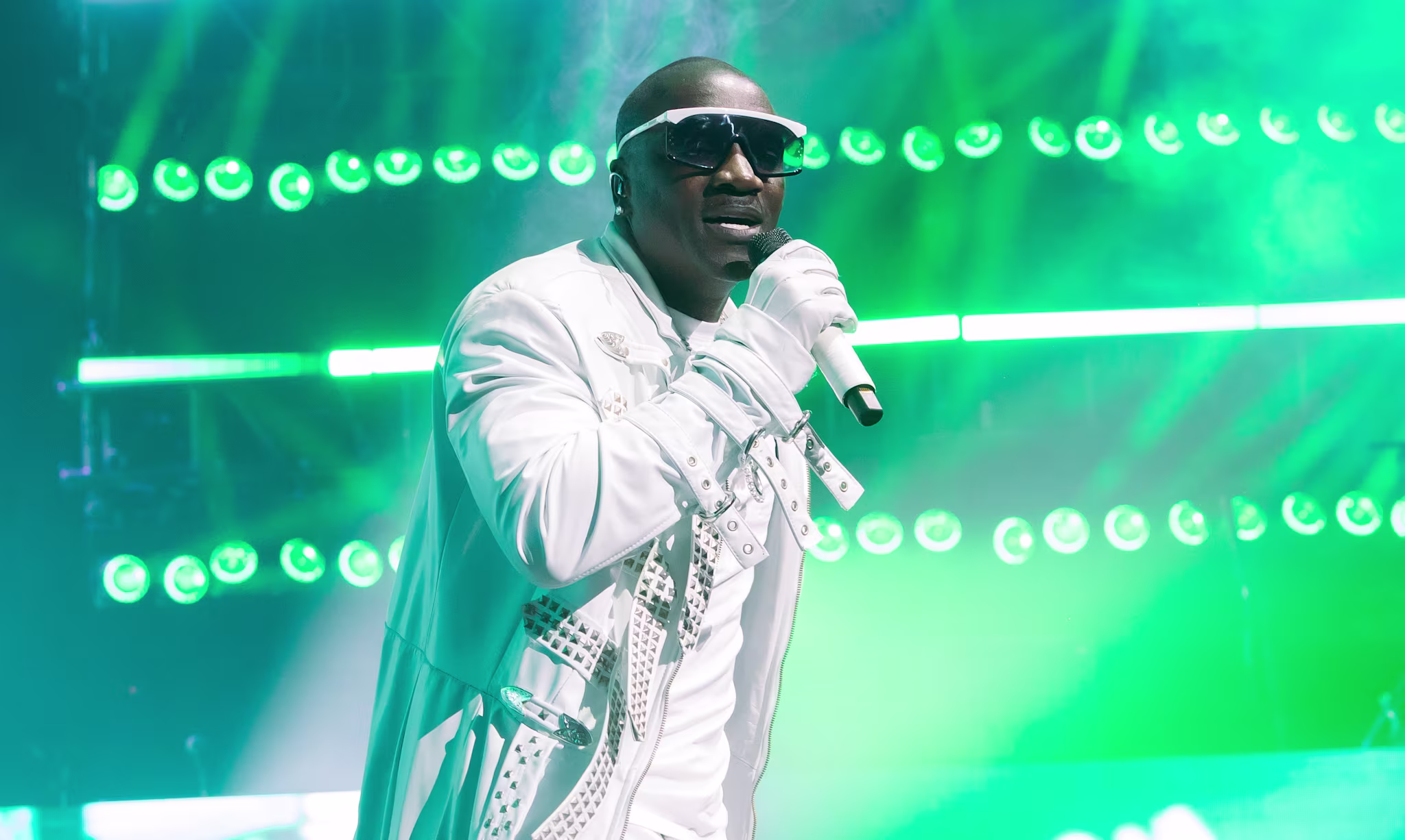 Akon in a white outfit and sunglasses sings on stage with vibrant green lights in the background.
