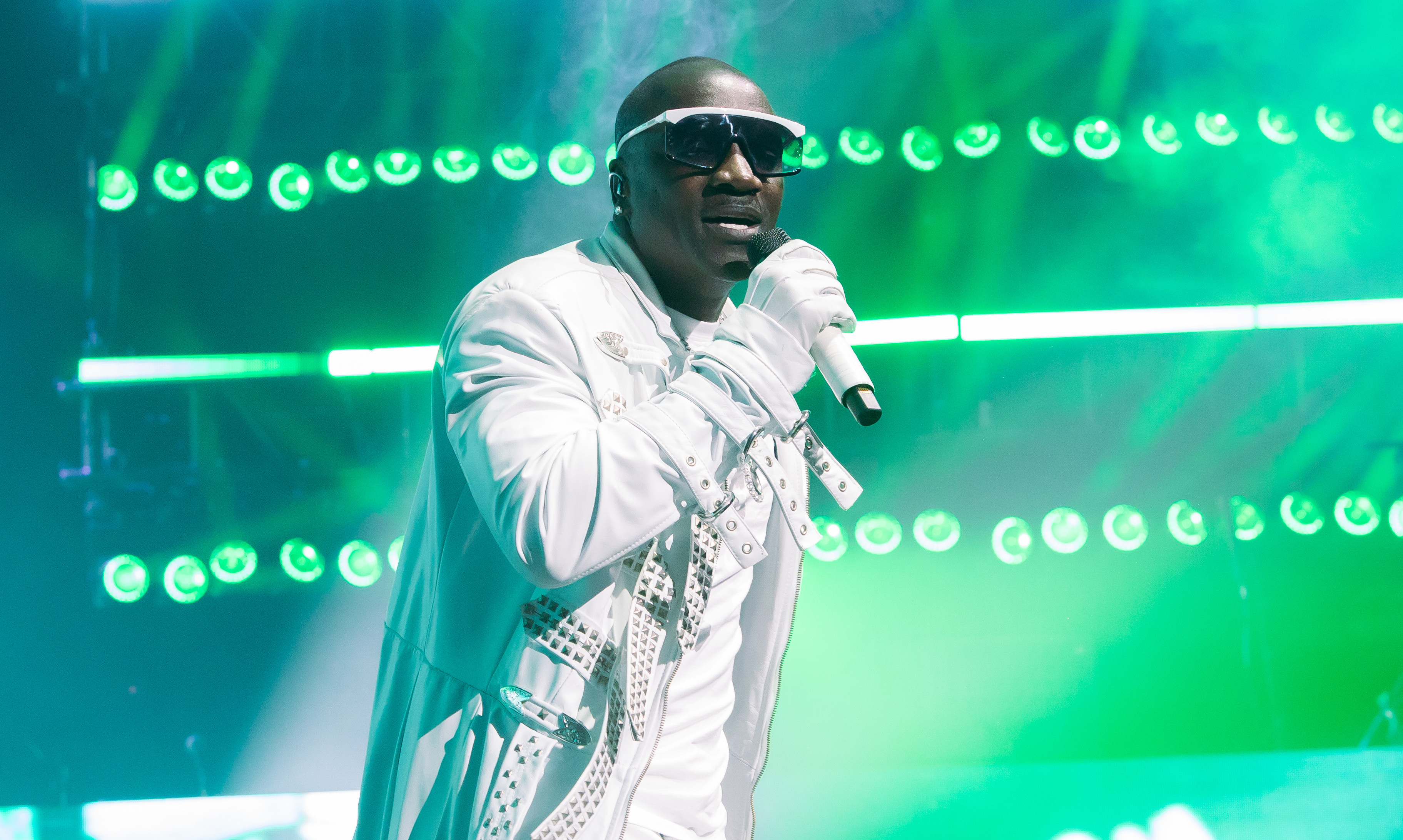 Akon in a white outfit and sunglasses sings on stage, surrounded by vibrant green lights and visual effects.