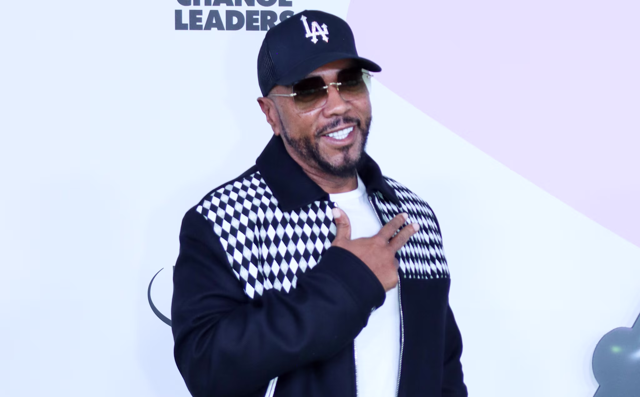 Timbaland in a black and white jacket, black cap, and sunglasses poses on a green carpet at an event.