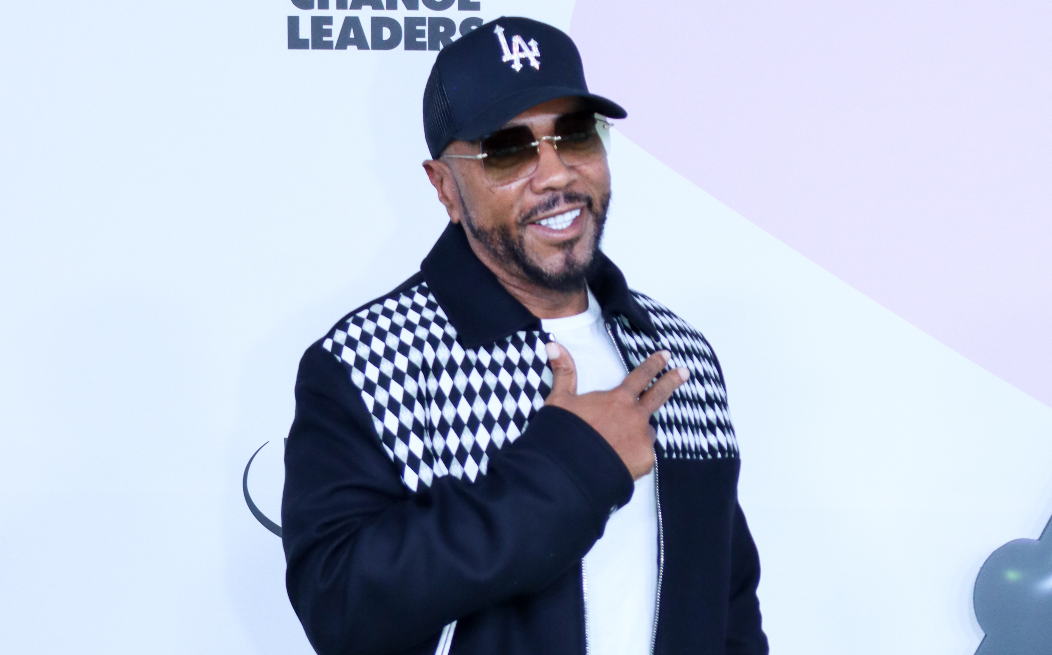Timbaland in a black and white jacket, black cap, and sunglasses poses on a green carpet at an event.