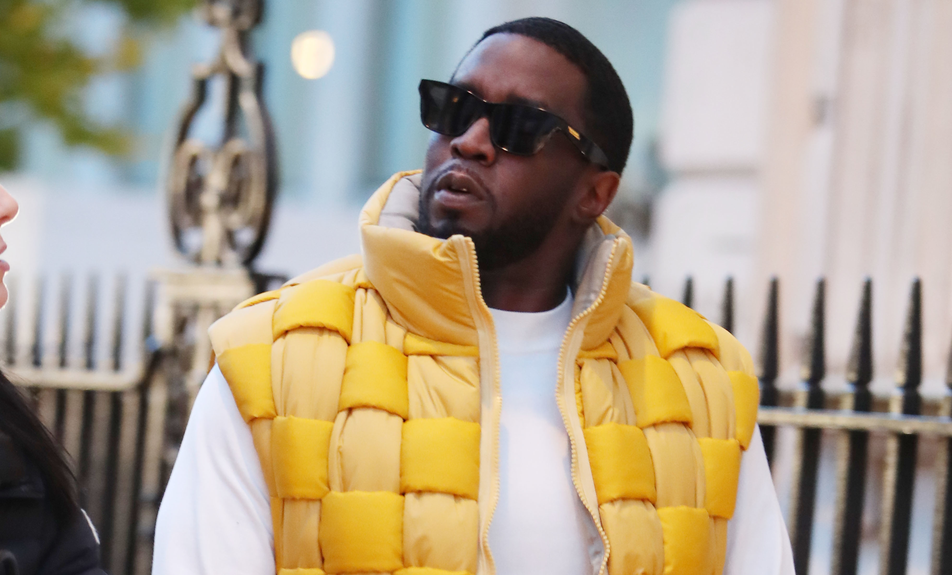 Diddy wearing sunglasses, a yellow puffer vest, white shirt, and jeans walks outdoors.