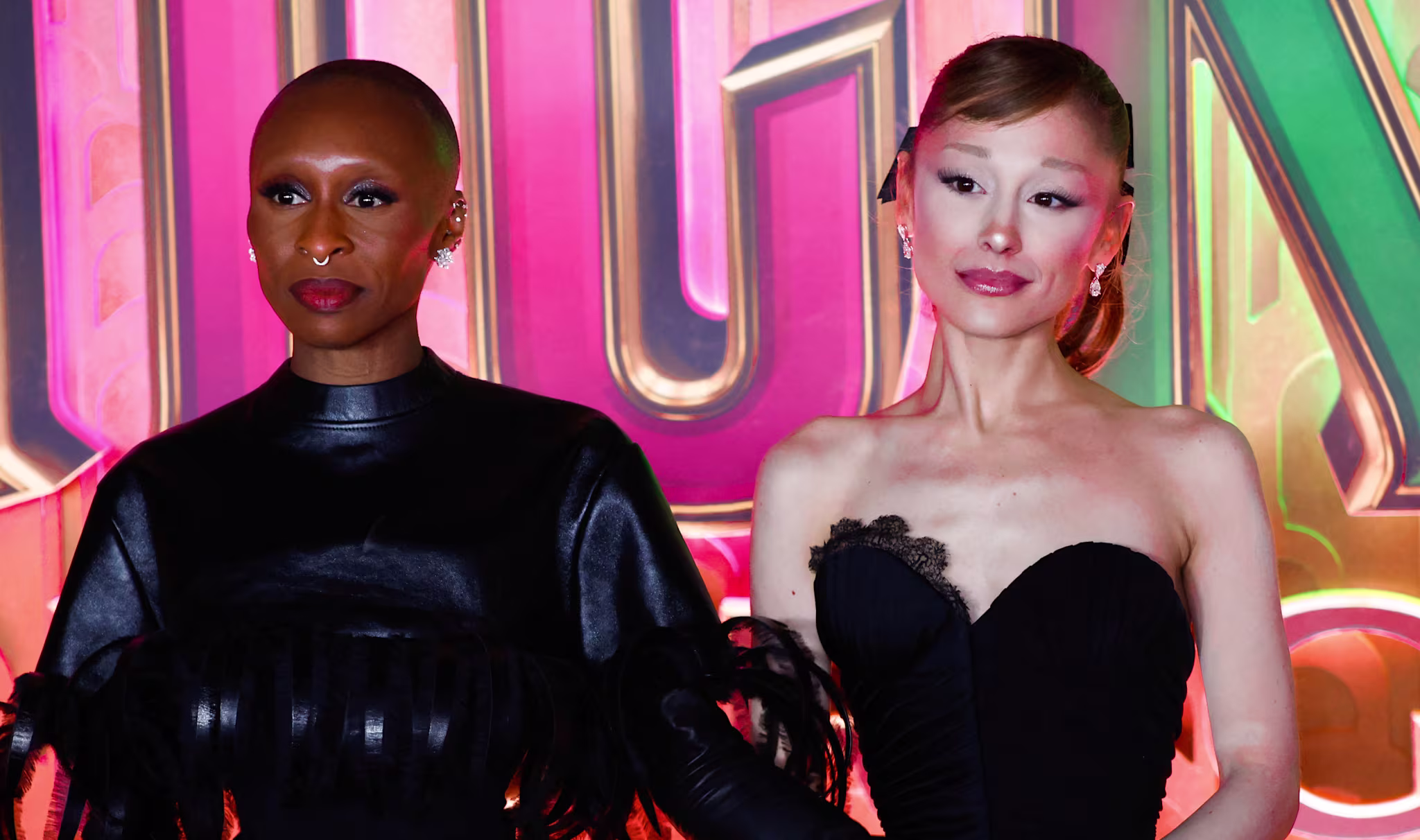 Cynthia Erivo and Ariana Grande pose together at an event, wearing elegant black outfits against a colorful backdrop.