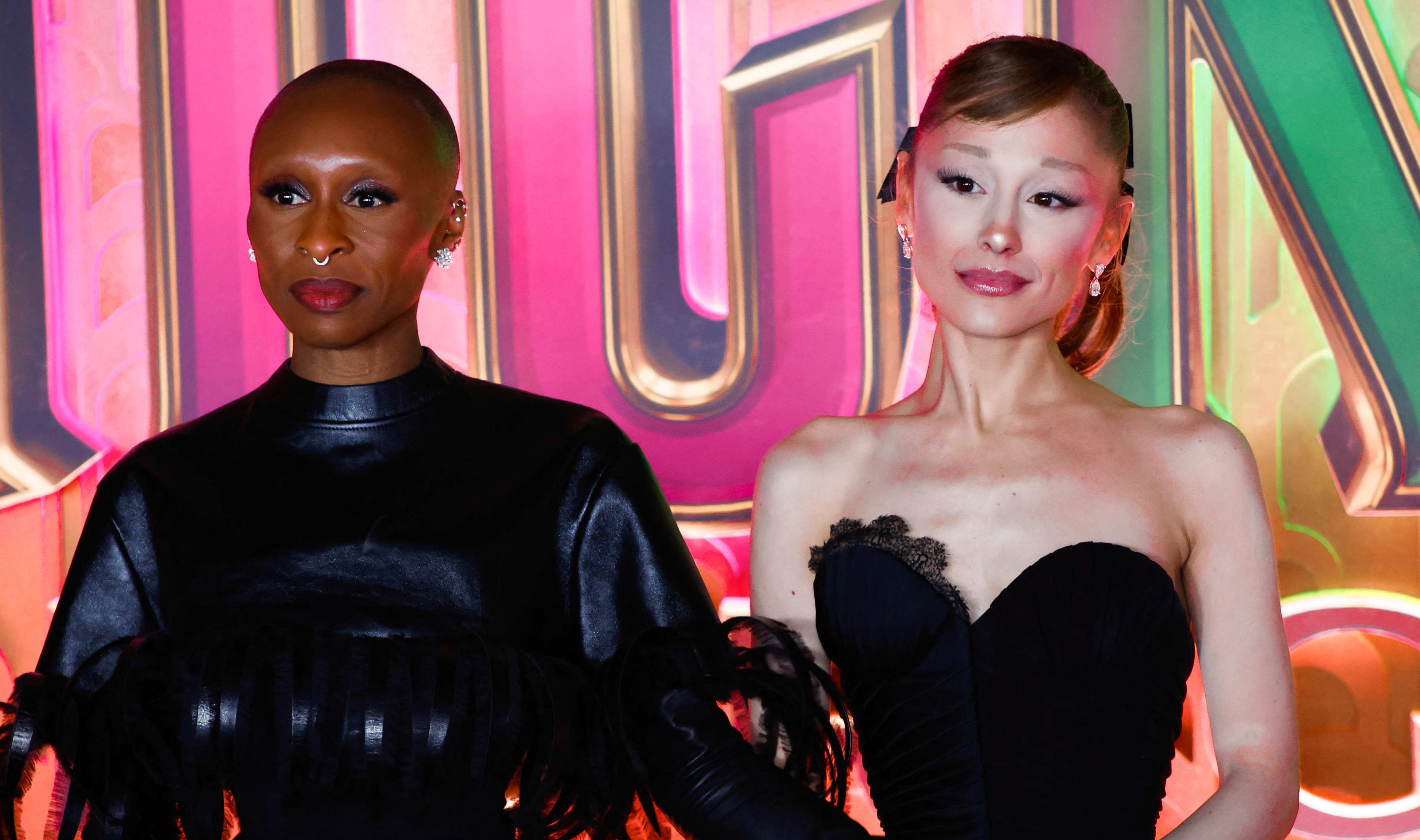 Cynthia Erivo and Ariana Grande pose together at an event, wearing elegant black outfits against a colorful backdrop.