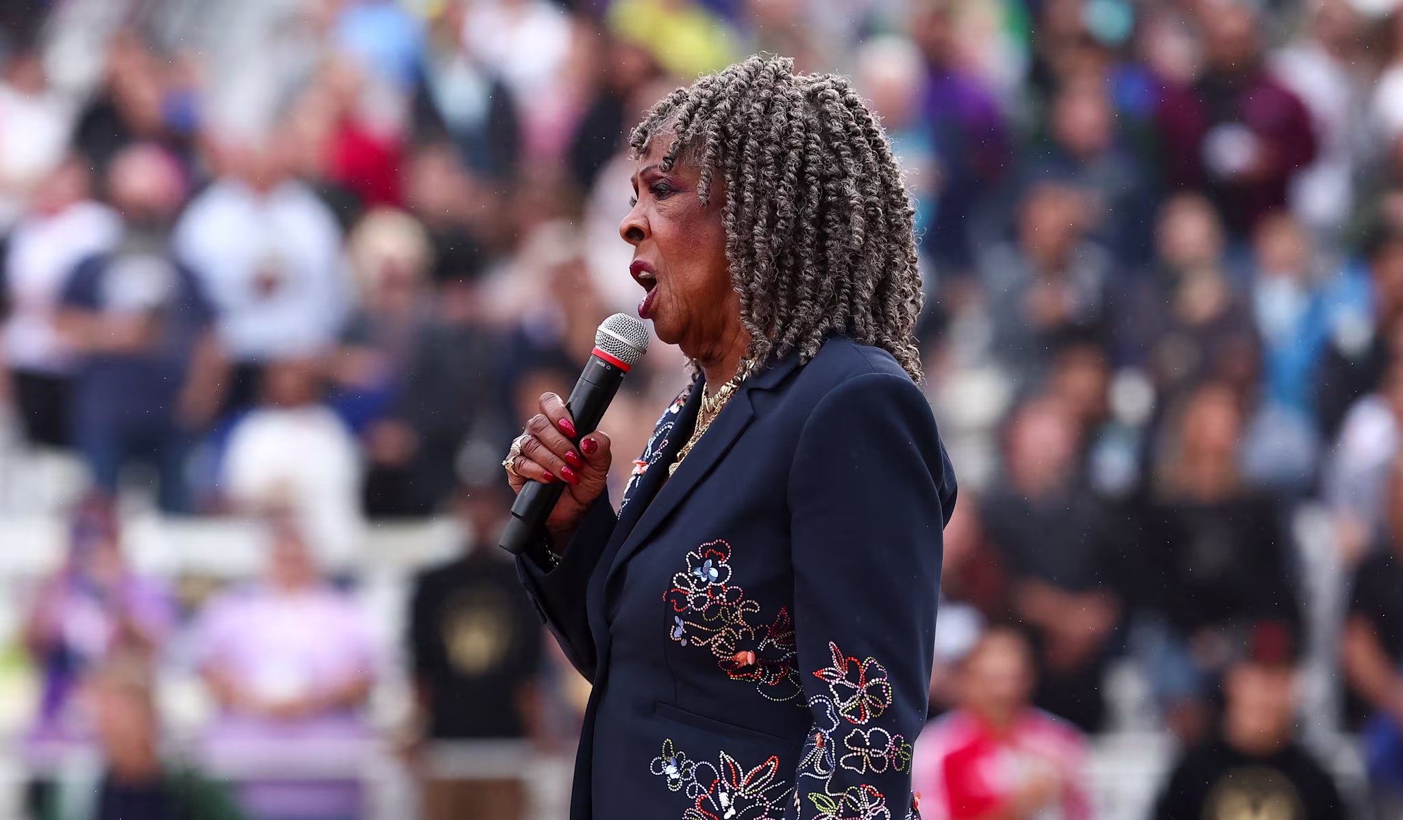 Martha Reeves with curly hair sings into a microphone at a stadium event, wearing a dark suit with floral embroidery.