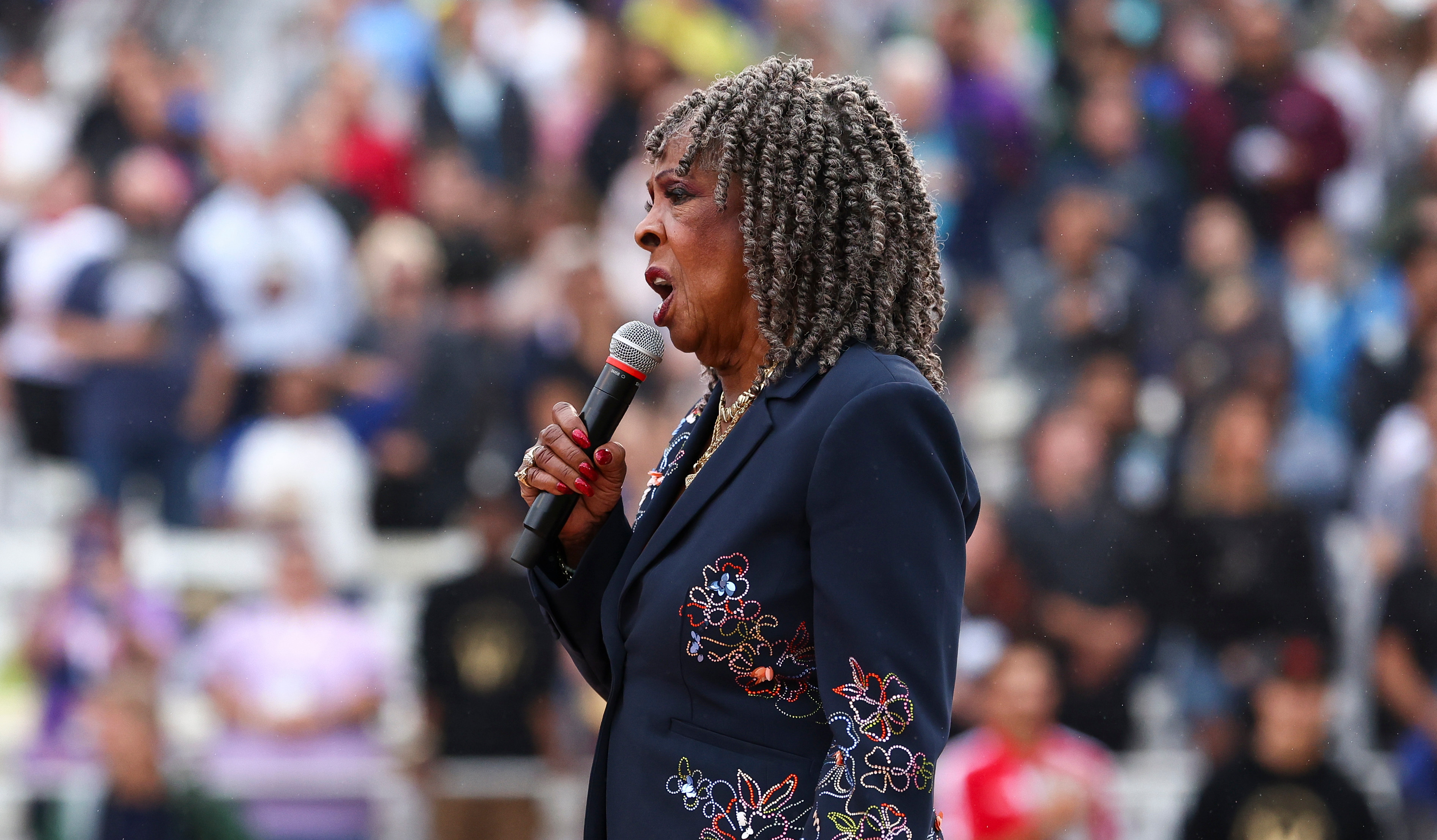 Martha Reeves with curly hair sings into a microphone at a stadium event, wearing a dark suit with floral embroidery.