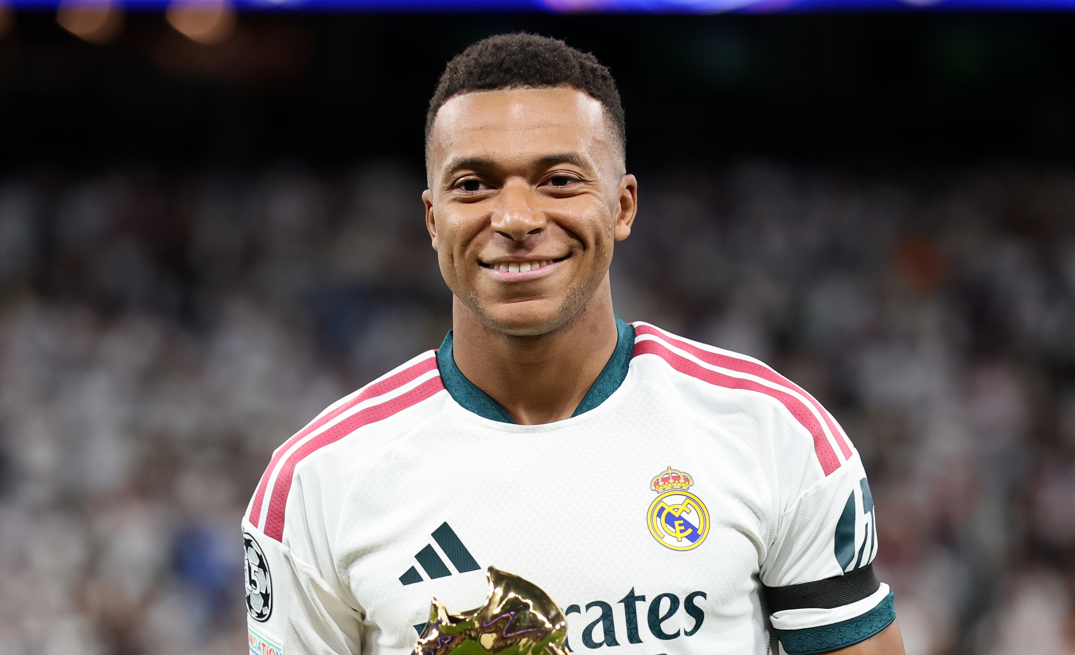 Kylian Mbappe in a Real Madrid jersey holds a golden trophy on a field, smiling. The background shows a blurred stadium crowd.