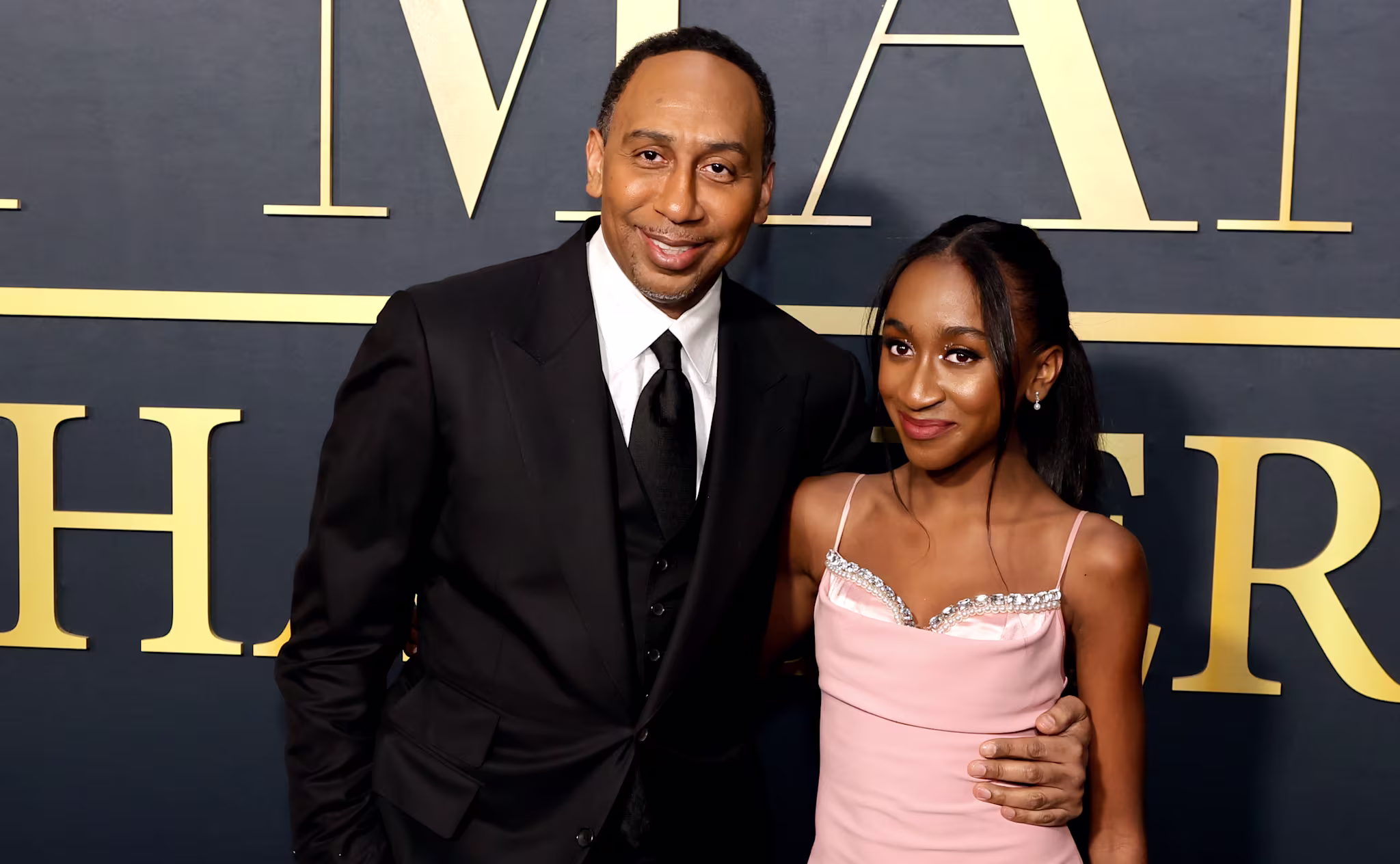 Stephen A. Smith in a black suit and his daughter in a pink dress pose together at an event with a dark backdrop.