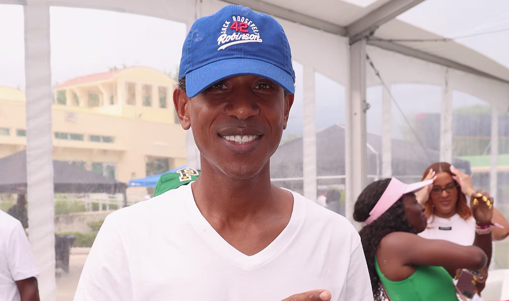 Shyne in a white shirt and blue cap is smiling and pointing in a casual indoor setting. People are in the background.