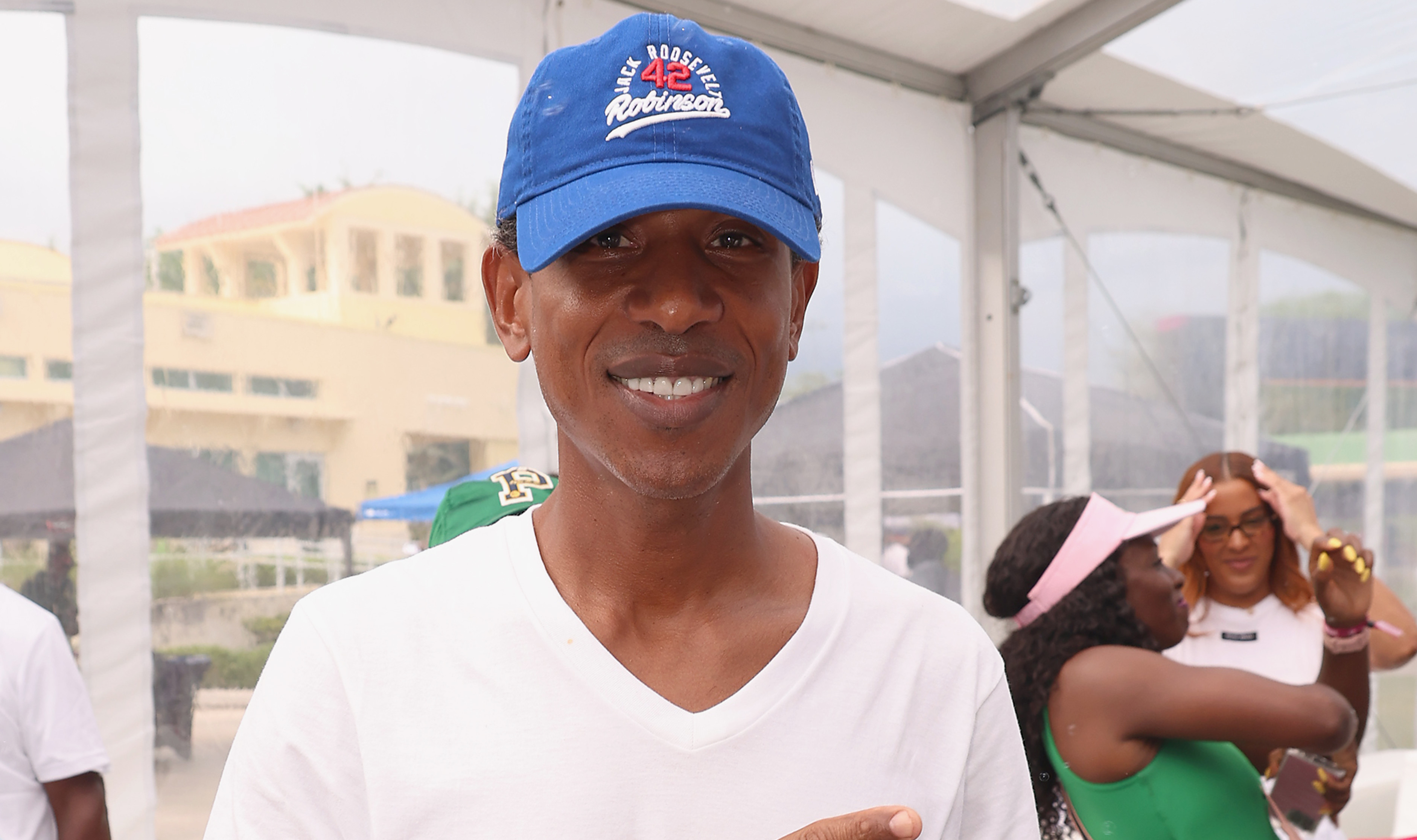 Shyne in a white shirt and blue cap smiling and pointing, with people in the background at an outdoor event.
