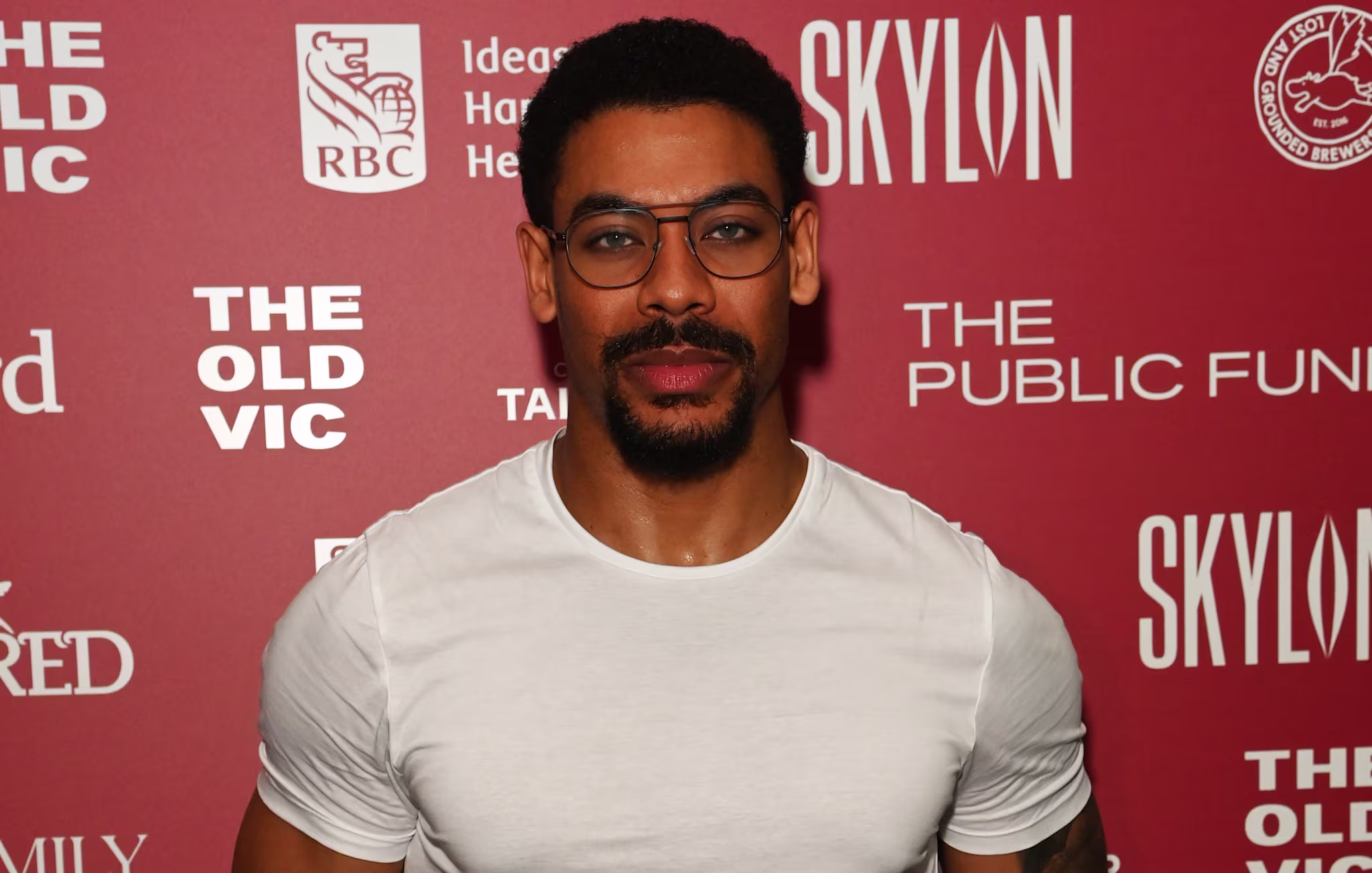 Aaron Pierre in a white shirt and glasses stands in front of a red backdrop with logos.