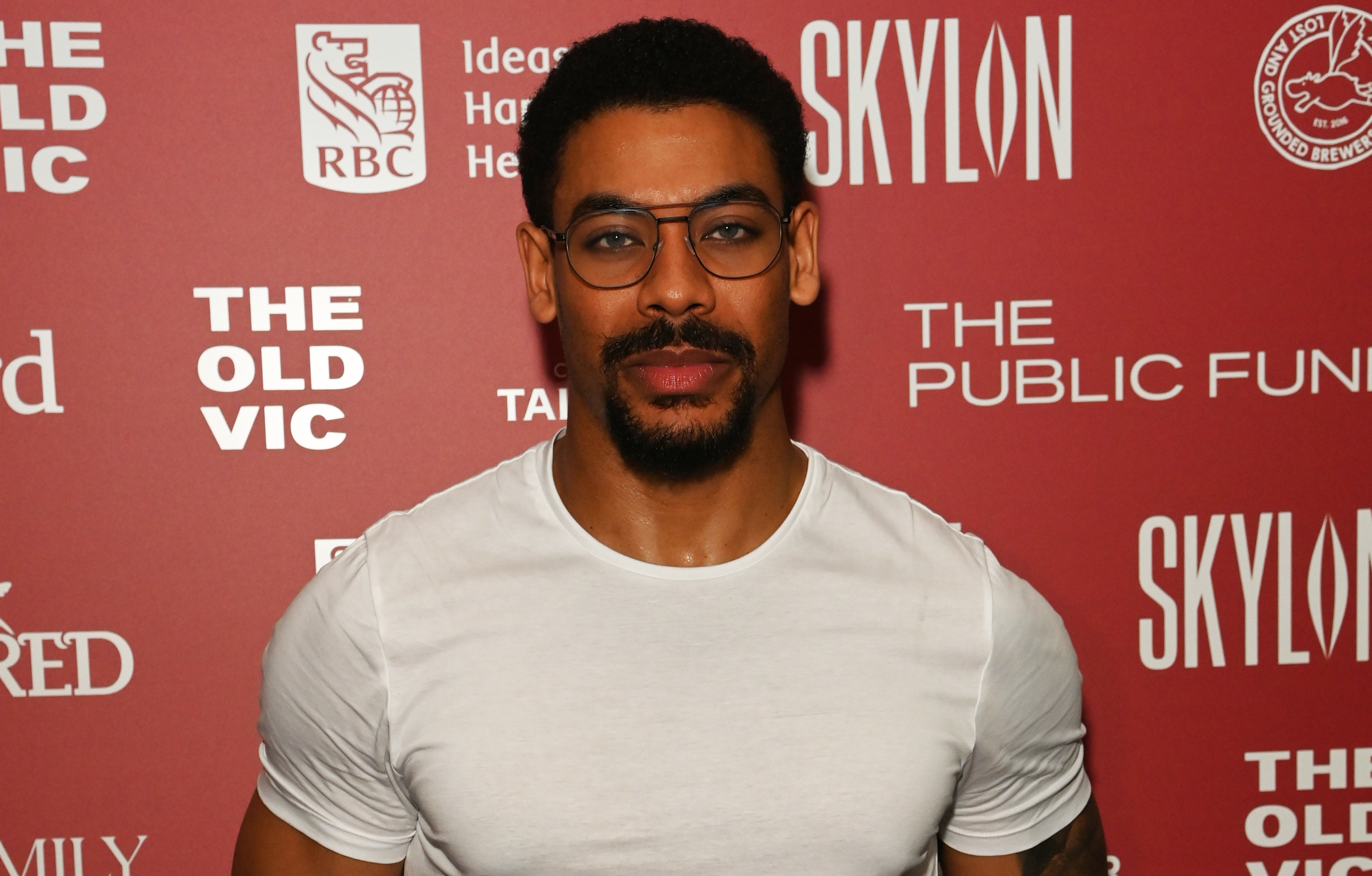 Aaron Pierre in a white shirt and glasses stands in front of a red backdrop with logos.