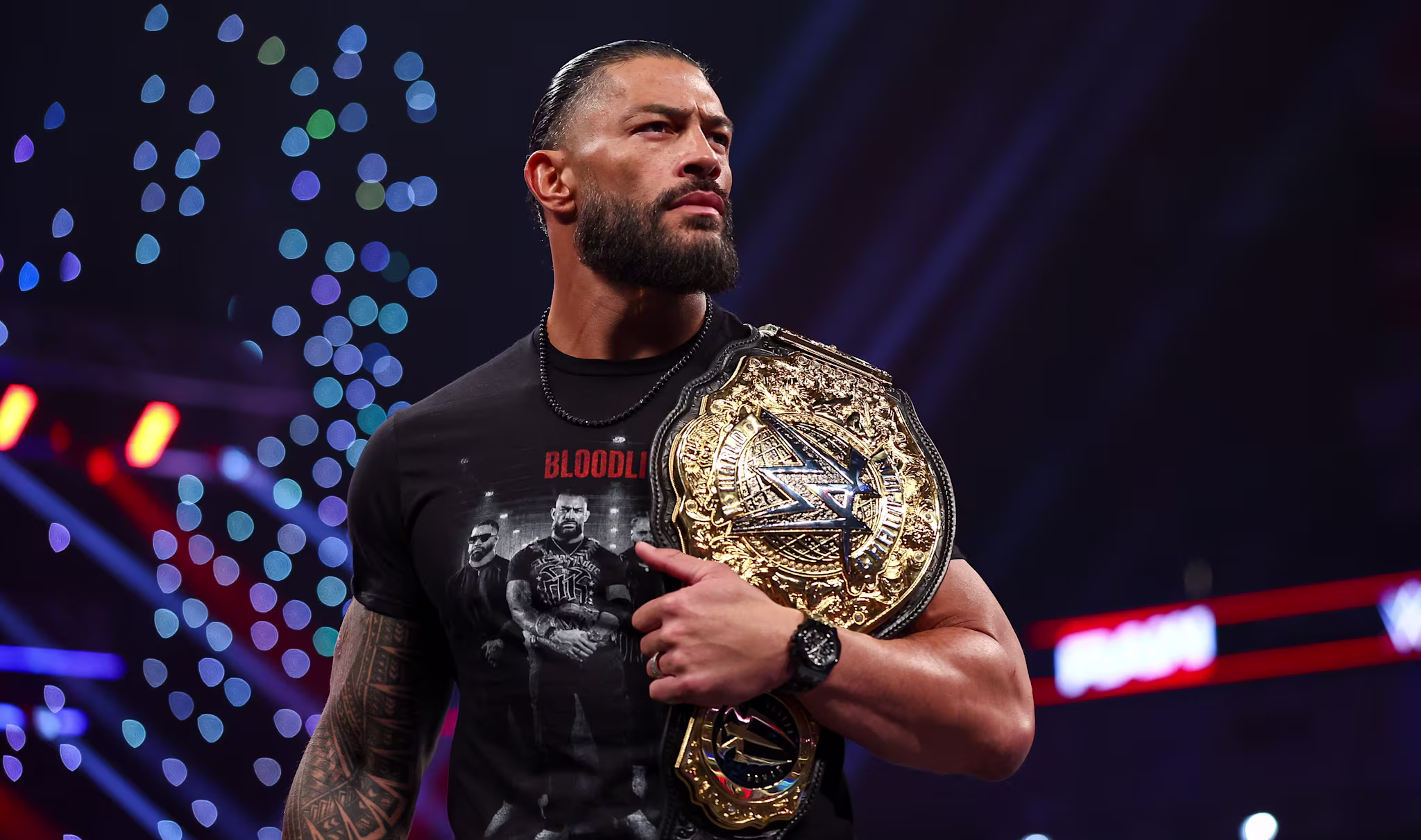Roman Reigns holding a championship belt, wearing a black T-shirt, with a focused expression. Lights and stage visible in the background.