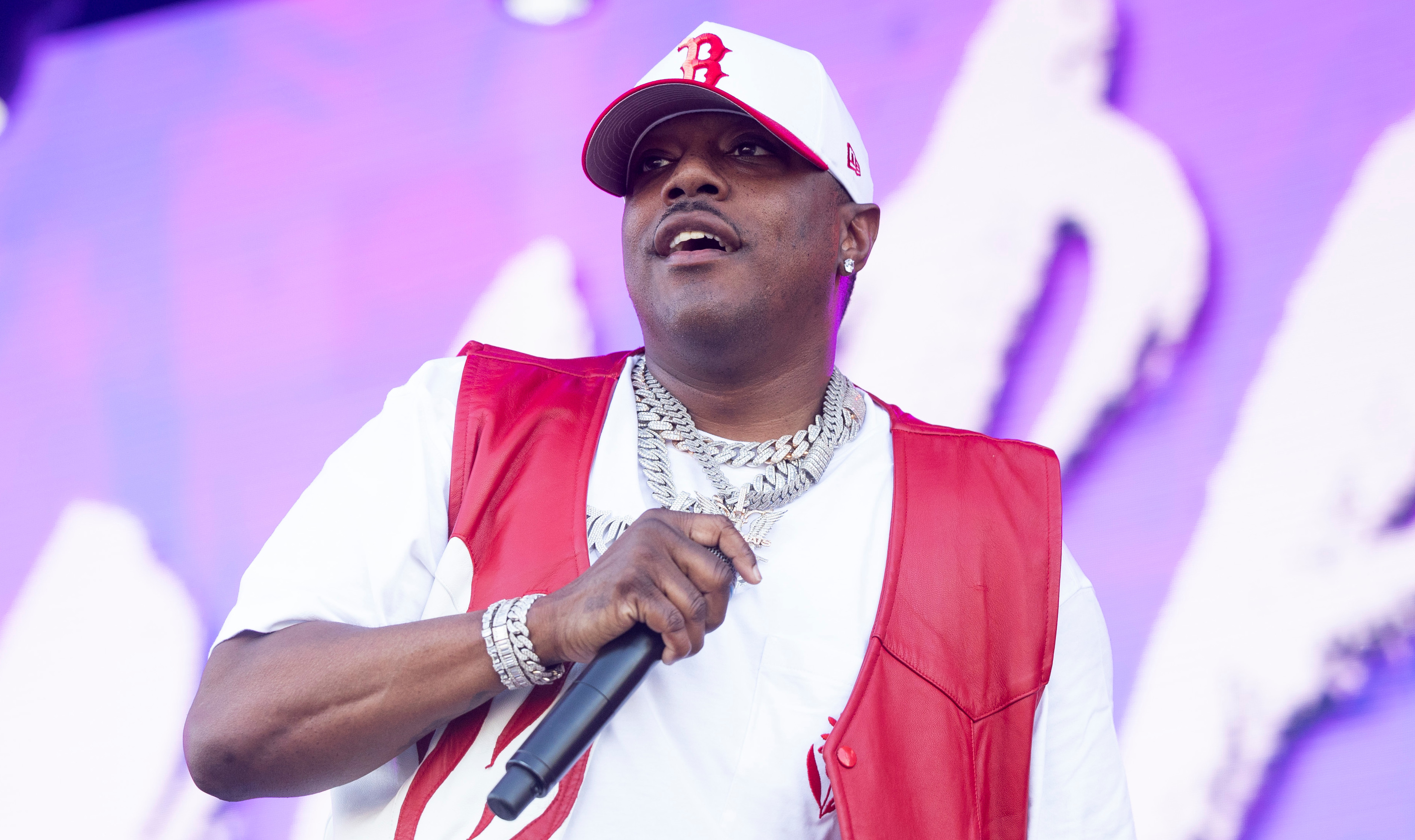 Ma$e performs on stage wearing a red and white outfit, holding a microphone, with a vibrant background.
