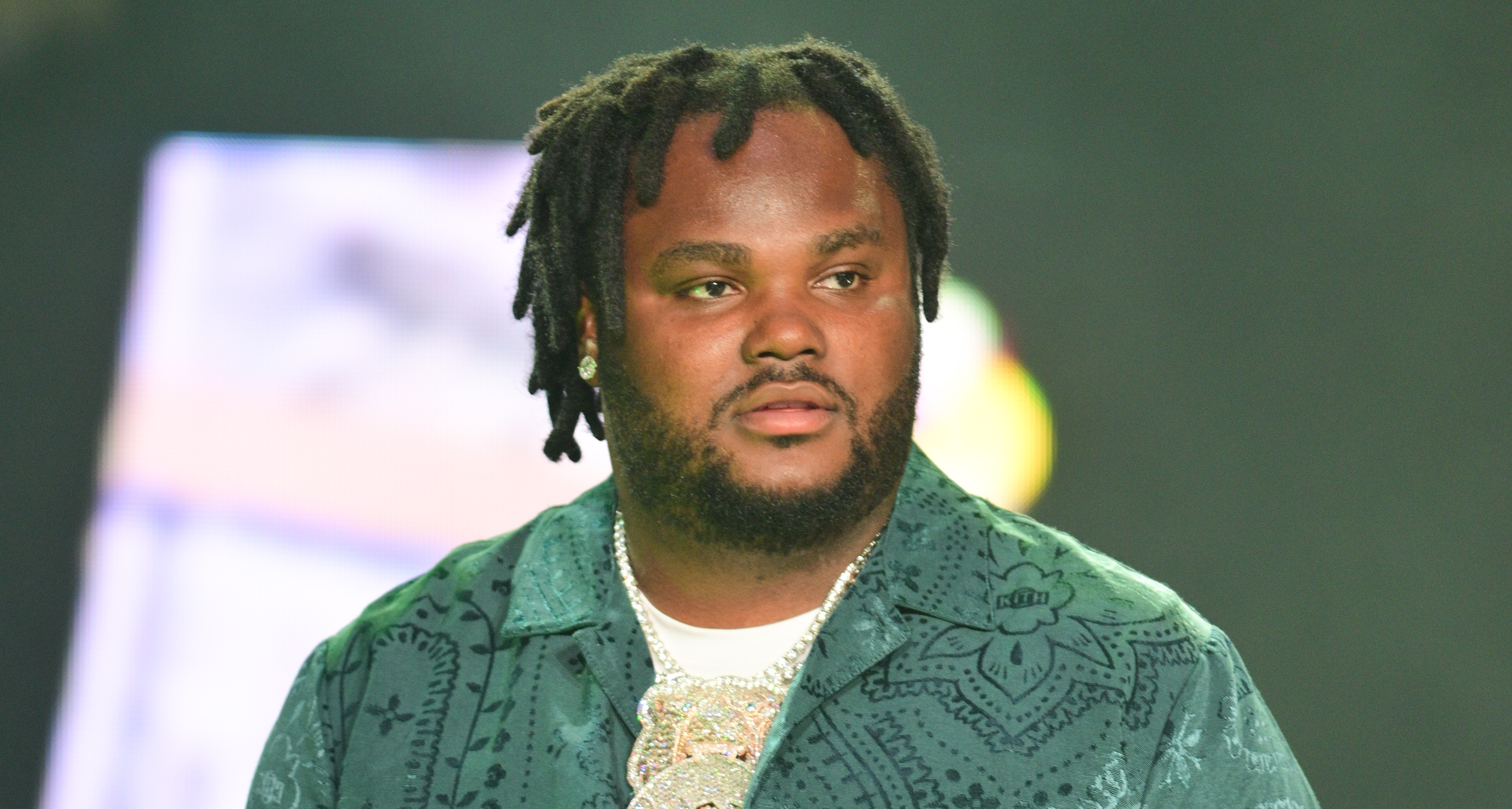 Tee Grizzley in a green patterned outfit stands on stage, holding a microphone, with a blurred background.