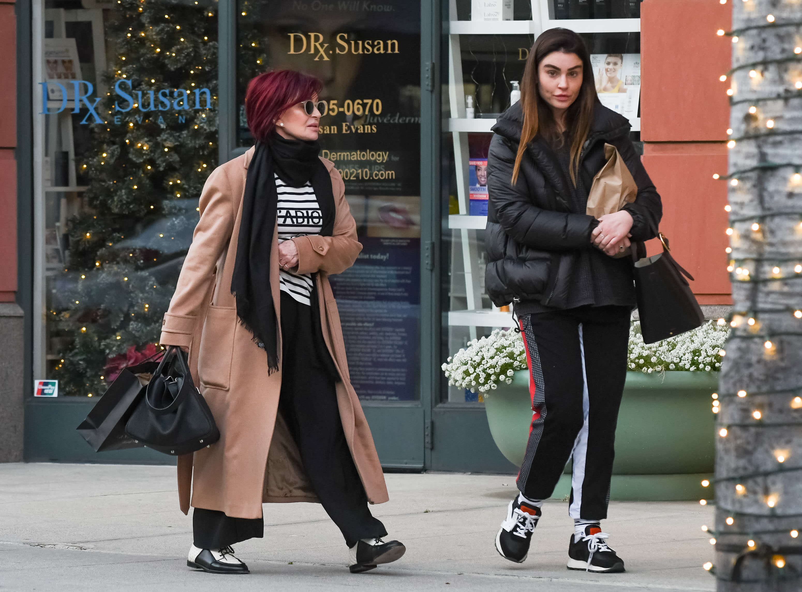 Sharon and Aimee Osbourne walking on a sidewalk, one with red hair in a long coat, the other in a black jacket, near a decorated storefront.