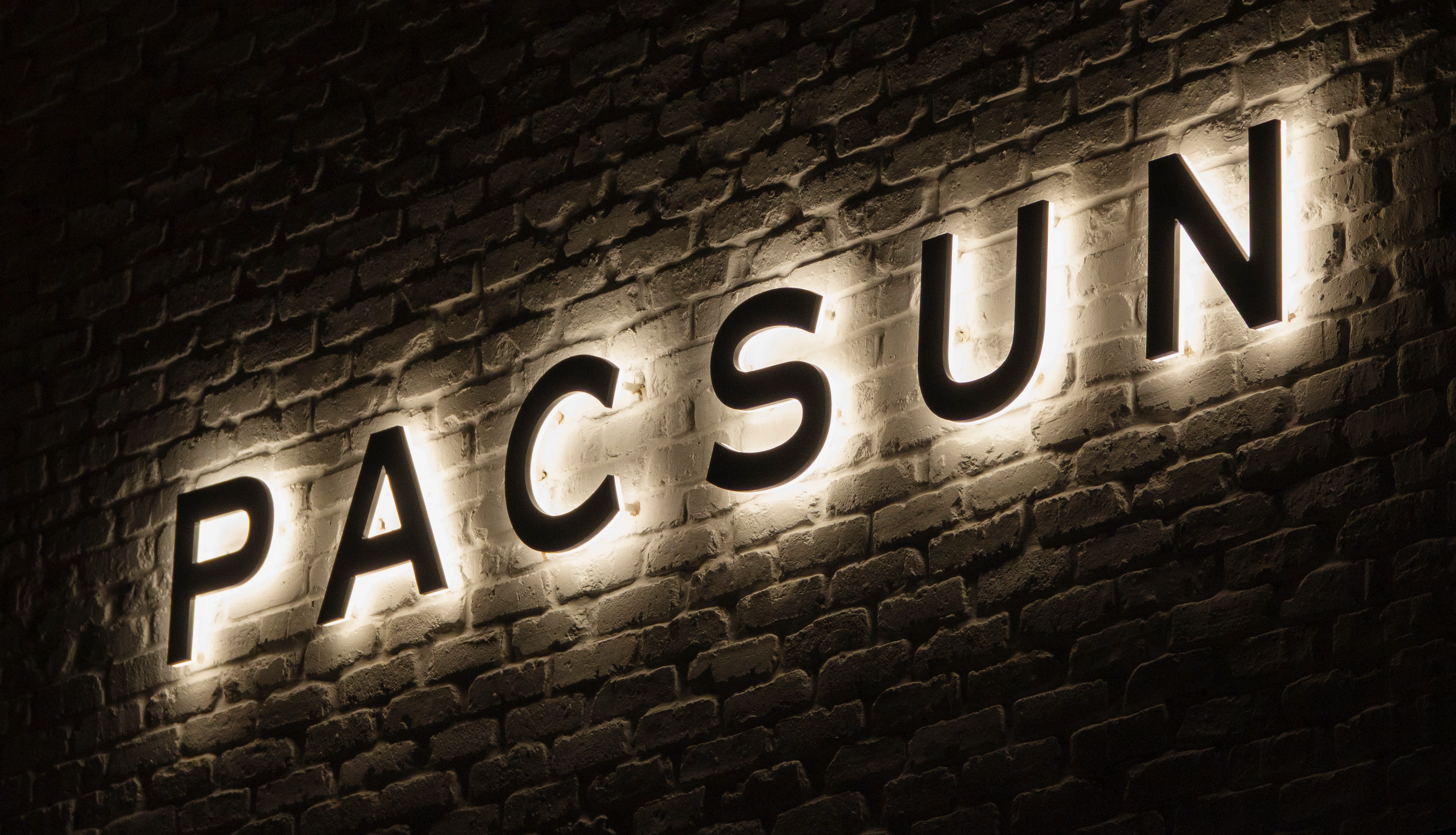 Illuminated "PACSUN" sign on a brick wall, glowing softly in the dark.