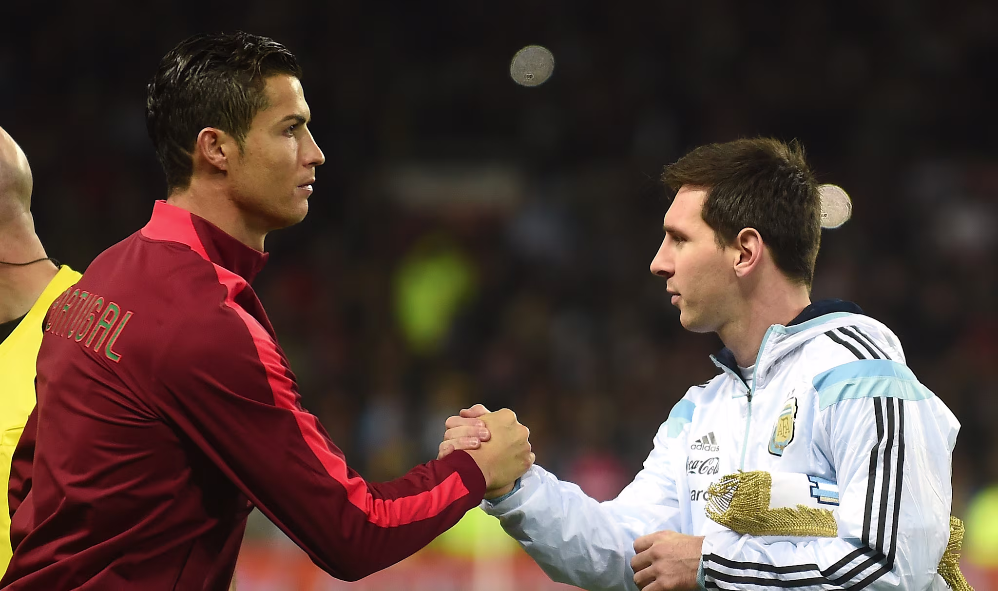 Cristiano Ronaldo and Lionel Messi shaking hands on a football field, wearing Portugal and Argentina jerseys respectively.
