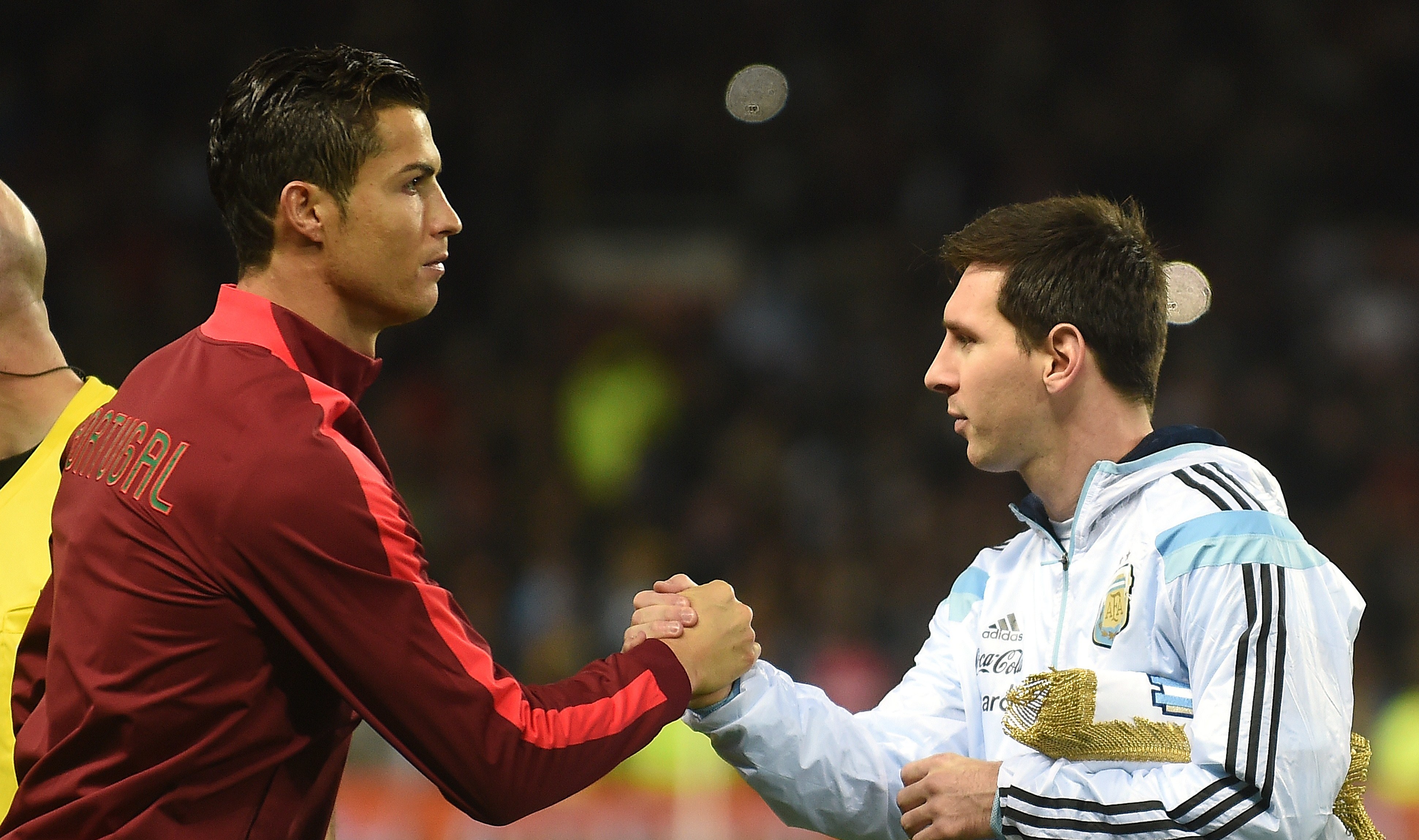 Cristiano Ronaldo and Lionel Messi shaking hands on a soccer field, both in their national team gear.