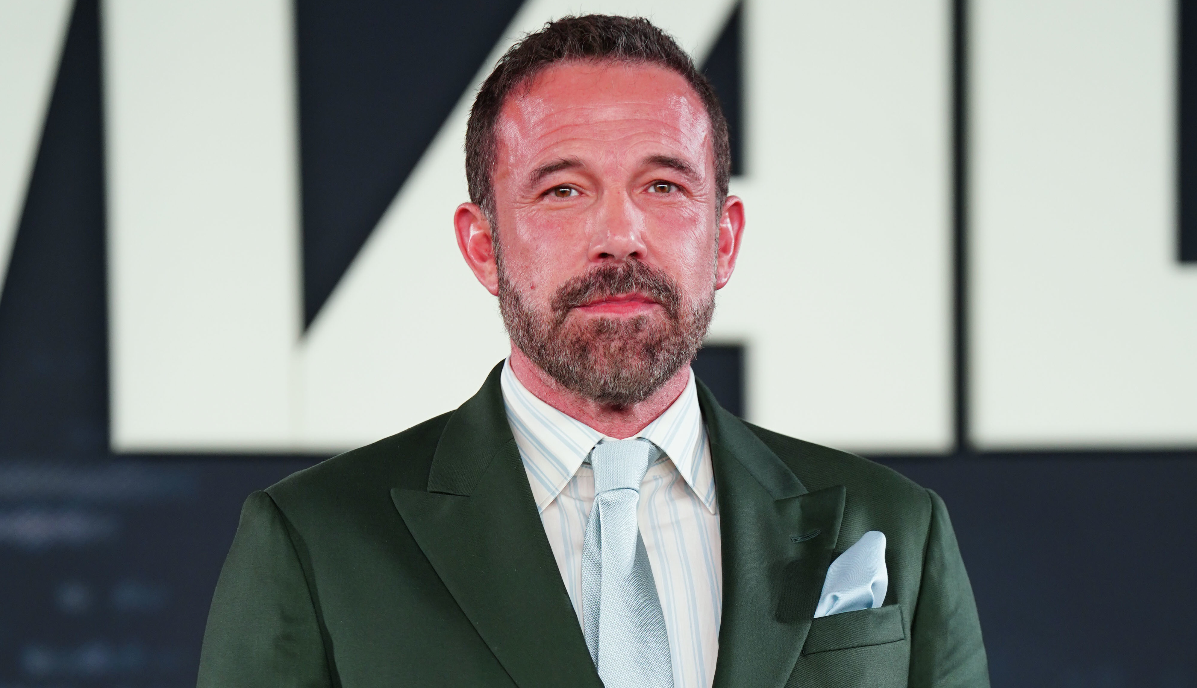 Ben Affleck in a green suit with a light blue tie and pocket square stands against a dark background.