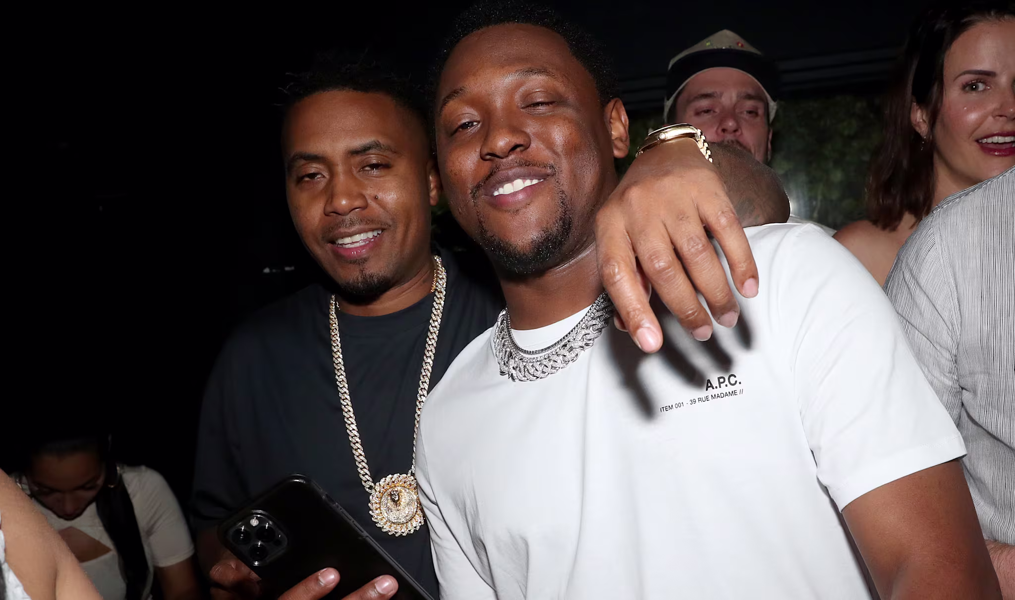 Nas, wearing a black shirt and gold chain, poses with Hit-Boy at an event. They are smiling and holding a phone.