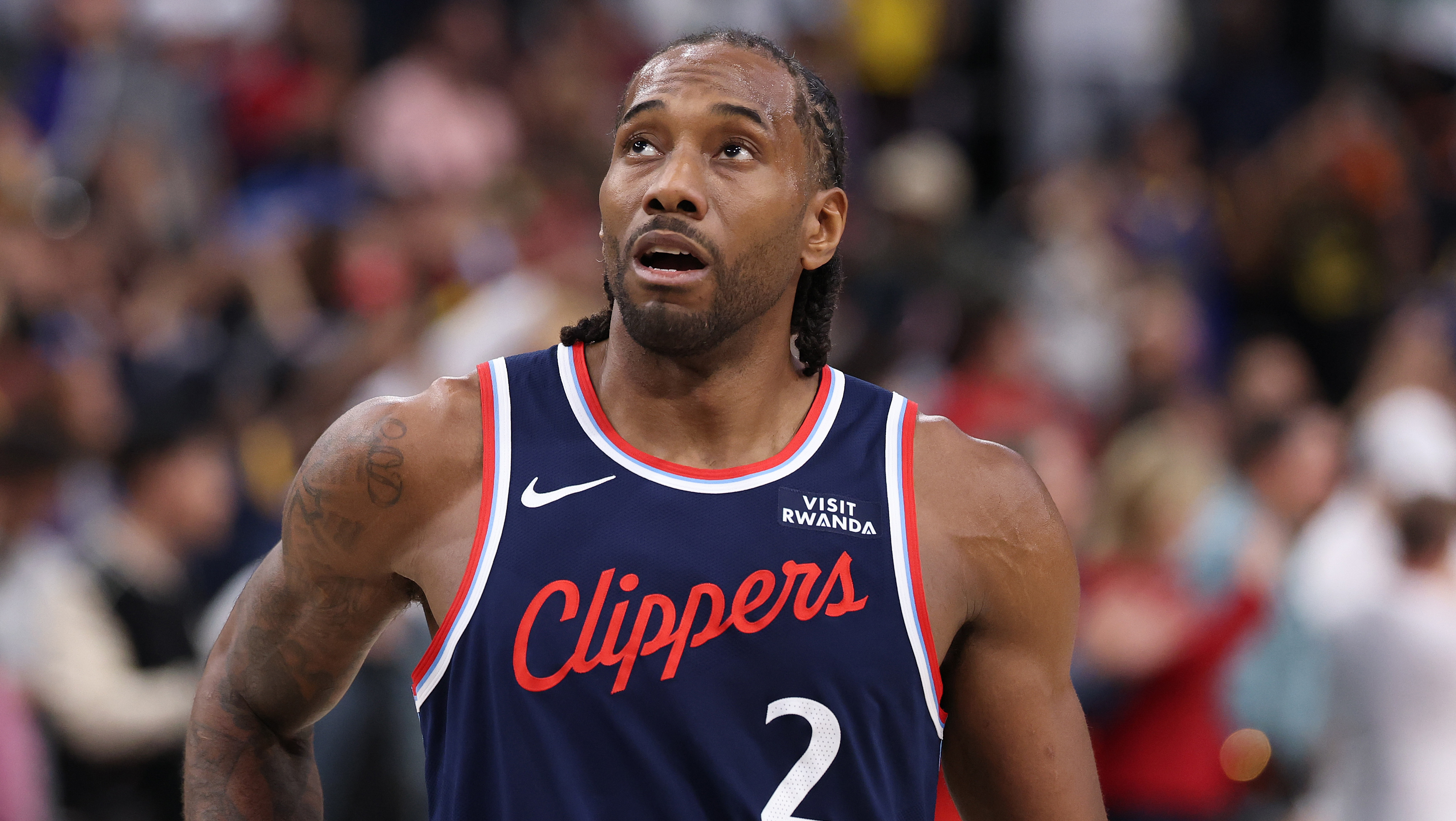 Kawhi Leonard in a Clippers jersey on the court, looking upwards, with a crowd in the background.