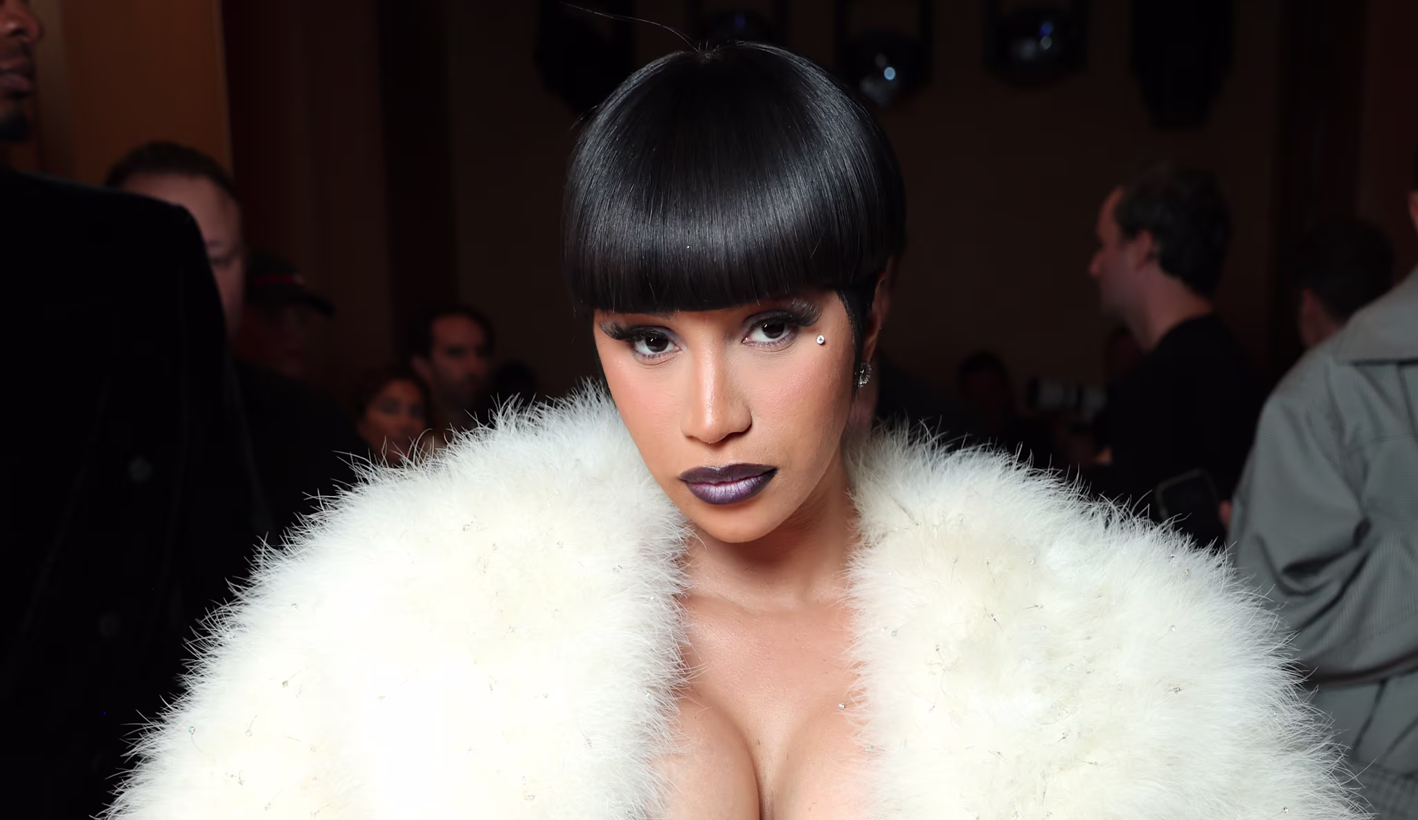 Cardi B with a sleek bob hairstyle wearing a luxurious white fur coat, standing in a dimly lit room with people in the background.
