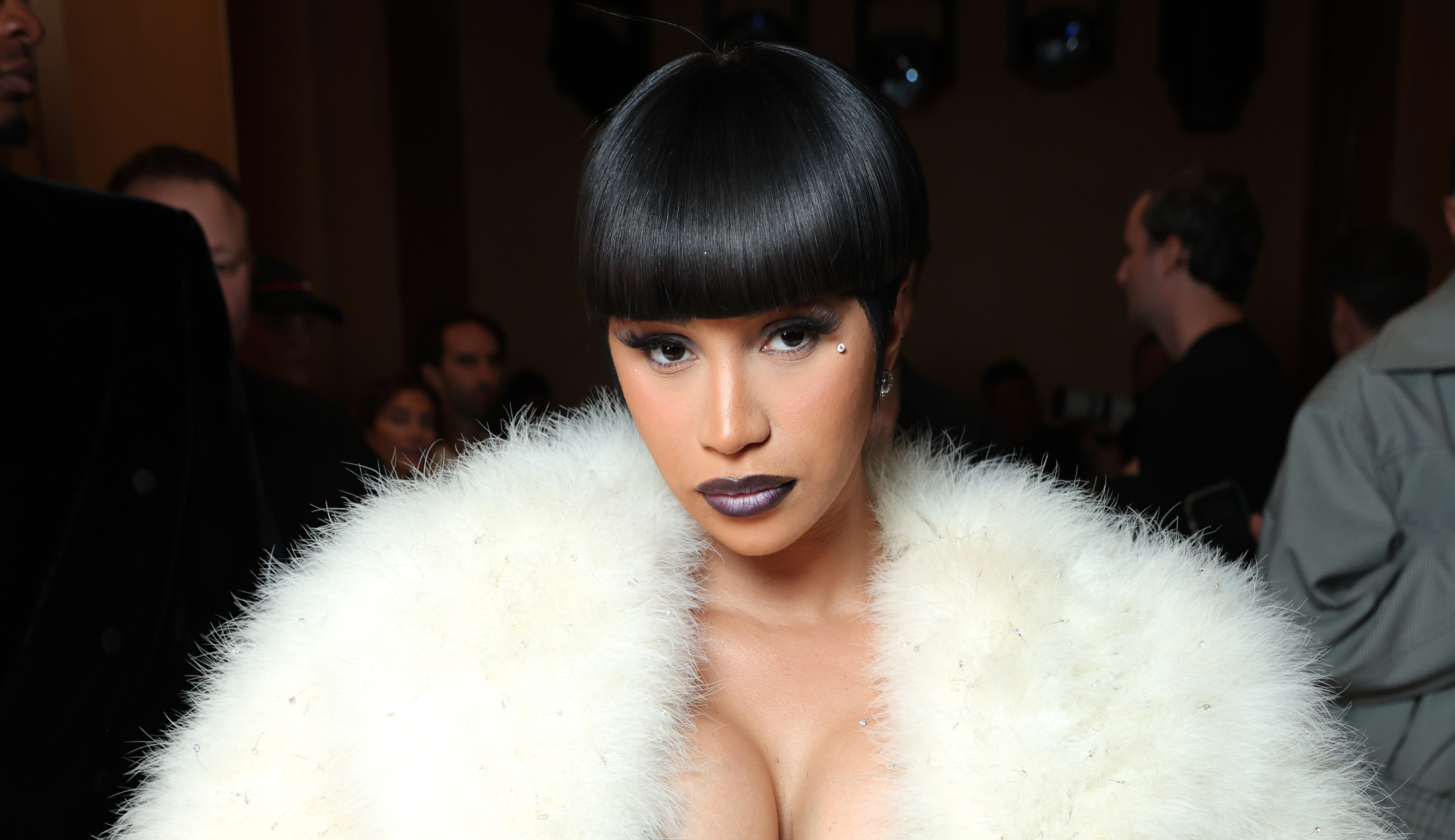Cardi B in a white fur coat and dark lipstick stands in a crowded room, looking directly at the camera.