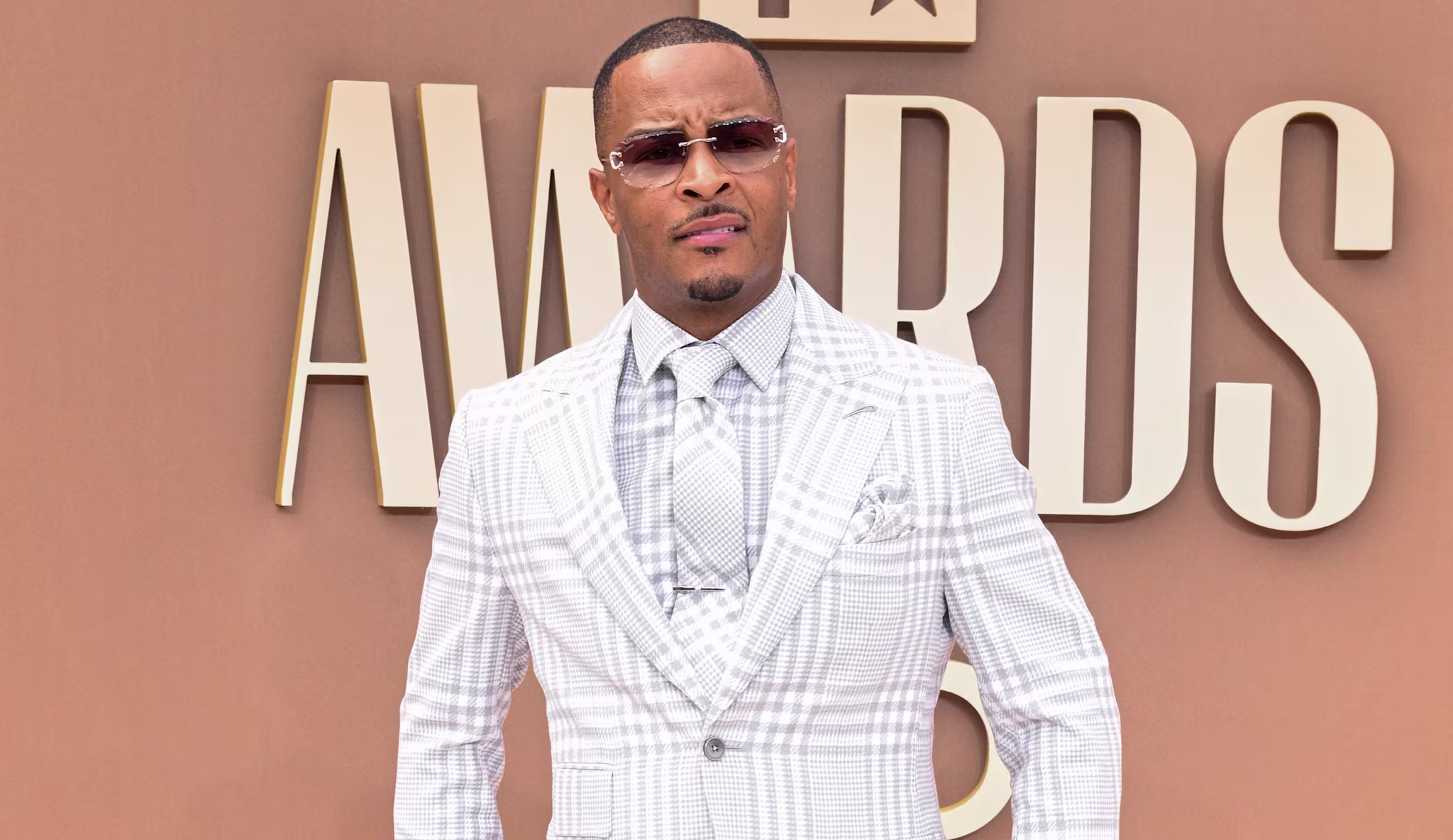 T.I. in a plaid suit and sunglasses stands on a red carpet at an awards event.